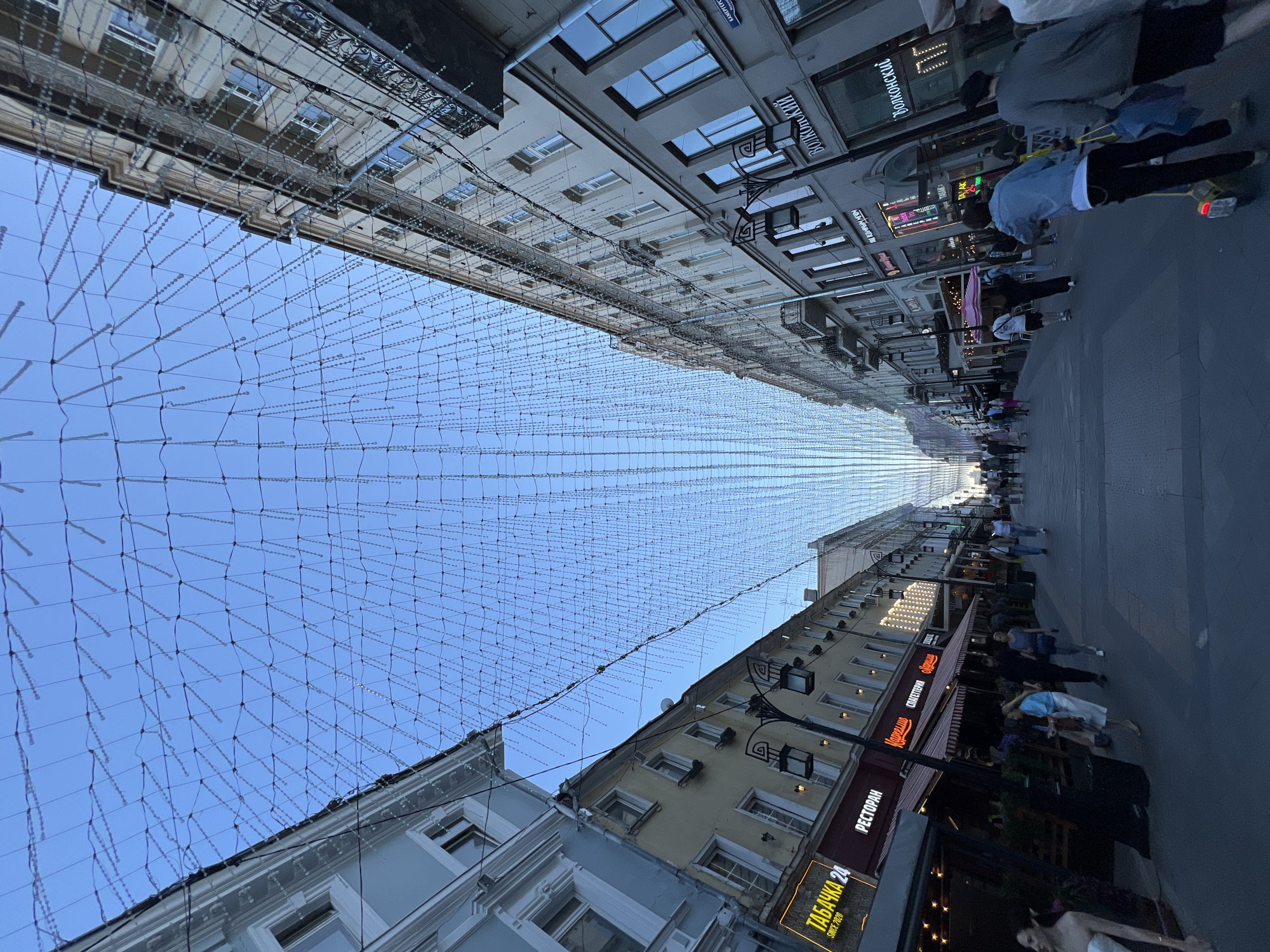 This image depicts Kamergersky Lane, a pedestrian street in Moscow, Russia. The scene is captured with a wide-angle lens, looking down the street, which is flanked by multi-story buildings. Overhead, an extensive canopy of decorative lights, appearing as a dense network of strings with small bulbs or beads, stretches across the entire width of the street, creating a tunnel effect that converges in the distance. The sky above the light installation is clear and blue.
On the left side of the street, buildings exhibit varying facades, including a light blue-grey structure in the foreground and a yellow building further back. Ground-level establishments include "ТАБАЧКА 24" (Tobacco 24), "РЕСТОРАН" (Restaurant), and "КАРЕЛЛИНО СПАГЕТТЕРИЯ" (Karellino Spagetteriya), with outdoor seating areas under striped awnings. A street lamp with two lanterns is visible on the left side.
On the right side, the buildings are predominantly light grey or beige, featuring multiple windows and balconies with black wrought-iron railings on upper floors. Ground-level businesses include "ВОЛКОНСКИЙ" (Volkonskiy), a bakery, and other shops. A street sign in Cyrillic for "КАМЕРГЕРСКИЙ ПЕР." (Kamergerskiy Per., i.e., Kamergersky Lane) is visible on a building facade.
Numerous pedestrians are present on the paved street, walking in both directions. Individuals are dressed in casual attire. In the left foreground, a woman in a white strapless dress is partially visible. Further down the street on the left, a woman in a light blue top and white skirt walks alongside a man in a dark shirt and a woman in a grey top. On the right side, a person on a bright yellow kick scooter is pushing what appears to be a blue bag, with another person in a grey shirt walking beside them. Other pedestrians are visible further into the distance, some partially obscured by one another or by street furniture. The overall lighting suggests either late afternoon or early morning, with long shadows cast across the street surface.