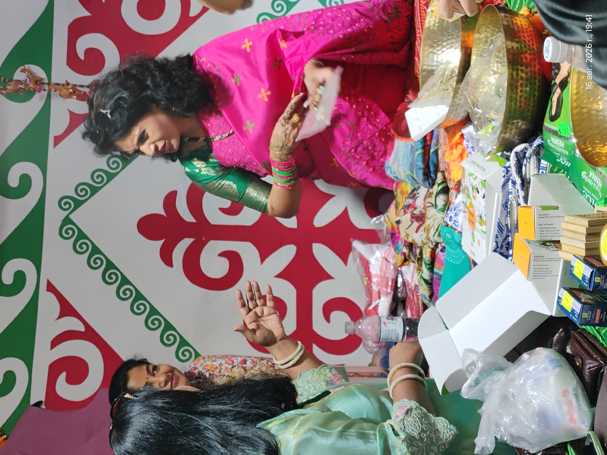 In Astana, Kazakhstan, an indoor scene unfolds during what appears to be a celebratory event, possibly a religious or cultural gathering. Three individuals are present. On the left, a woman with long, dark hair is partially visible, wearing a light green, shimmering outfit adorned with intricate embroidery on the sleeves and collar. She has several bangles on her wrists. To her right, another woman, also with dark hair, is smiling and looking towards the first woman. She is wearing a colorful, patterned blouse. The third individual, a woman with dark, curly hair, is the central focus. She is dressed in a vibrant pink sari with green trim and small embroidered motifs. She is wearing numerous bangles and a beaded necklace. Her right hand is raised, holding a small, clear packet and a gold ring is on her finger, with henna decorations visible on her hand. The backdrop is a large, decorative cloth with a striking pattern of red, green, and white swirls and geometric shapes. In the foreground, a table is laden with various items. These include several gift boxes, a plastic bag containing white objects, numerous colorful fabrics, a bottle of water, and two ornate brass bowls filled with what appear to be more wrapped items. The overall impression is one of a lively and possibly ritualistic occasion.