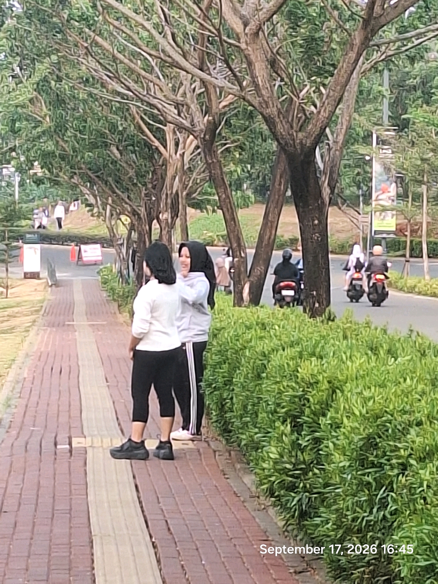 In Depok, Indonesia, two women stand on a brick-paved walkway bordering a lush green hedge. The woman on the left, wearing a white long-sleeved shirt and black leggings, faces away from the camera with her hands behind her back. The woman on the right, in a grey hoodie and black leggings with white stripes, faces the woman on the left, with her right hand touching the back of the other woman's head. A road runs parallel to the walkway, with several motorcycles visible. On the far left, a few people walk in the distance. The scene is framed by mature trees with bare branches, creating a dappled effect with the green foliage. The image is dated "September 17, 2026 16:45."