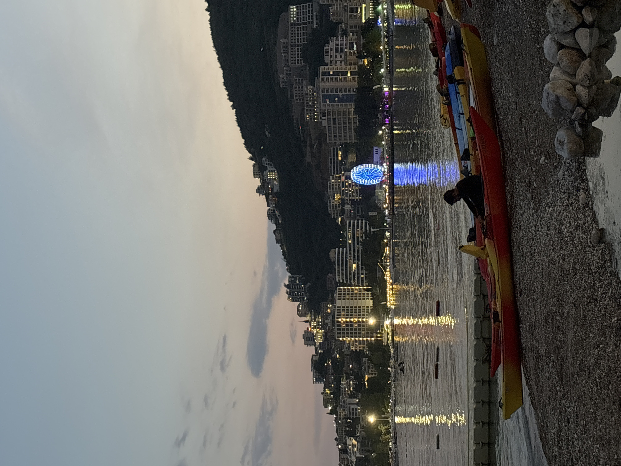 This image depicts the Budva Riviera in Montenegro at dusk. The scene is set on a pebble beach with several kayaks lined up. In the foreground, a person is seated in a red kayak. Beyond the kayaks, the calm sea reflects the lights of the city and a brightly lit Ferris wheel, which is a prominent landmark on the Budva waterfront. The background features a hillside densely populated with buildings, many of which are illuminated, creating a vibrant cityscape against the darkening sky. The overall atmosphere is serene and picturesque, characteristic of a coastal resort town in the evening.