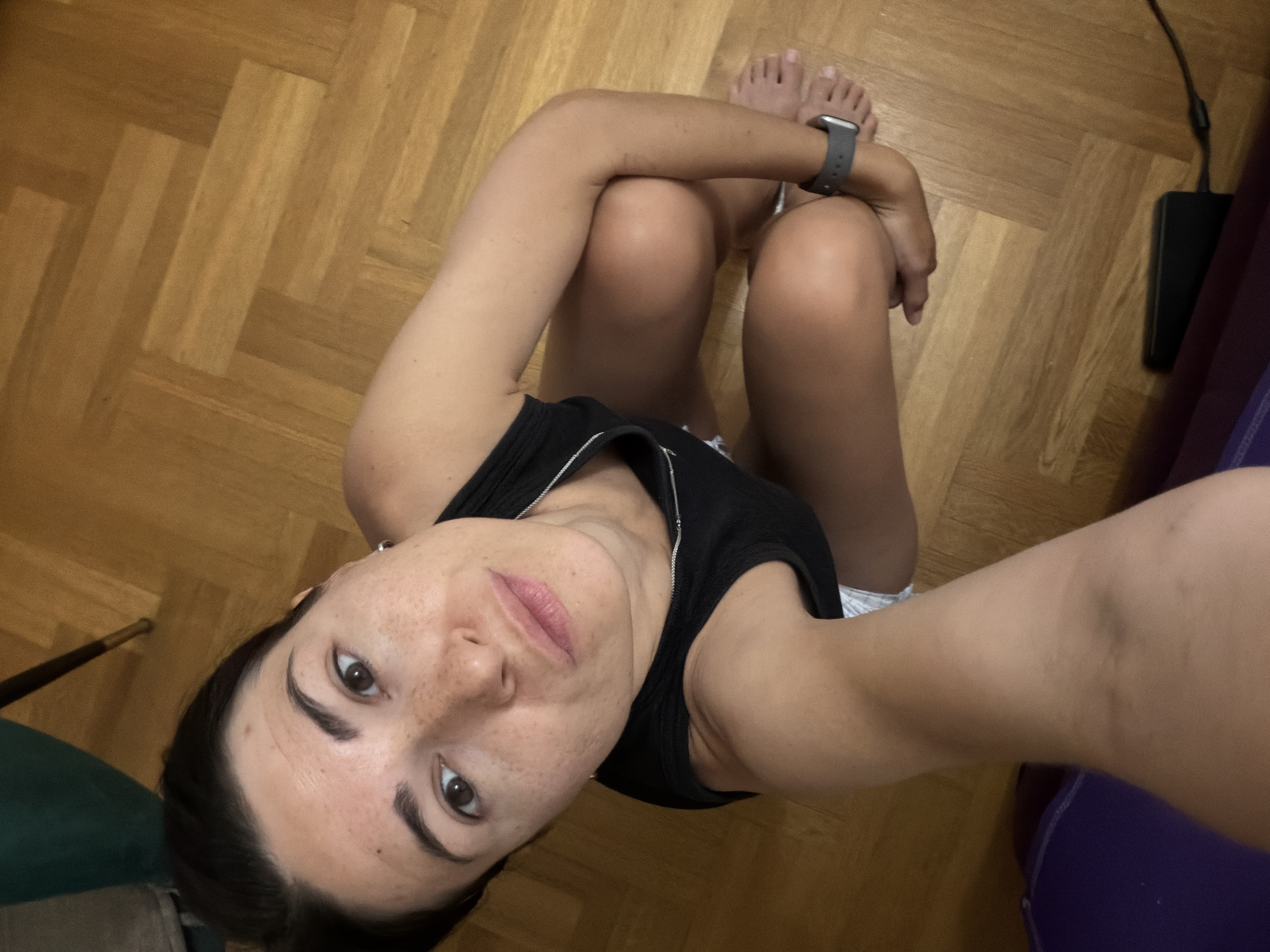 A woman with dark hair and fair skin is seated on a parquet floor. She wears a black tank top and a watch on her left wrist. Her legs are bent, and her arms are wrapped around them. She is looking up at the camera. The background includes a power adapter, a purple yoga mat, and a green object in the corner. The location is unknown. There is no indication that this image was taken in Budva, Montenegro.