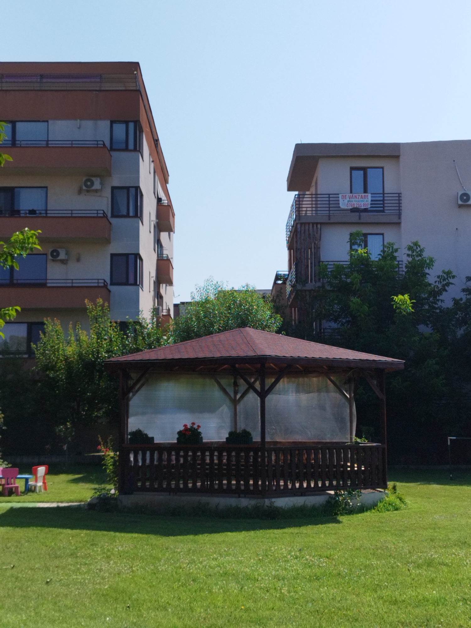 This is a medium shot, taken on a clear, sunny day, of a residential courtyard area. On the left, a tall apartment building with a reddish-brown facade on its upper levels and grey lower levels is visible. It has multiple balconies with dark railings. An air conditioning unit is mounted on the grey section of the building. To the right of this building is a smaller, lighter-colored apartment building, also with balconies. A "DE VANZARE" (For Sale) sign is attached to the railing of an upper balcony. Trees and dense green foliage are visible between and behind the buildings. In the foreground, a wooden gazebo with a dark brown roof and railing is situated on a concrete base. The sides of the gazebo are covered with transparent plastic sheeting. Inside the gazebo, a table and chairs are partially visible. In front of the gazebo, on the green lawn, are two small brightly colored plastic children's chairs and a blue table. The lawn is well-maintained. The scene appears to be a residential complex in Bucharest, Romania.