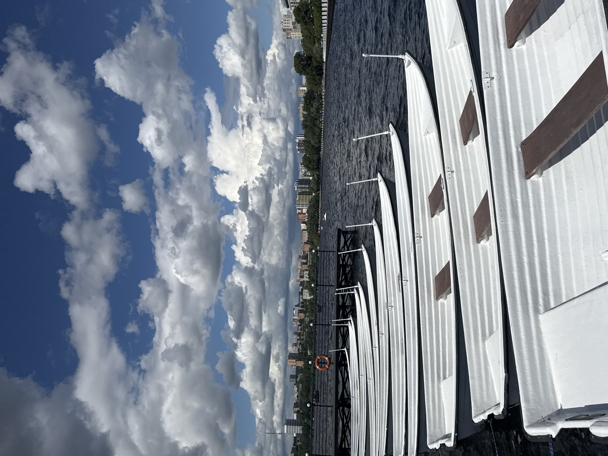 From a high-angle perspective, the image showcases a row of narrow white boats docked along a pier on a body of water, possibly the Izhevsk Pond, given the presence of what appears to be the city skyline of Izhevsk, Russia, in the background. The boats, characterized by their slender frames, are uniformly aligned, and each has a small brown seat or fixture. A thin metal bar separates the boats, acting as a support and keeping them evenly spaced. The water, dark and textured, reflects the sky. A black pier runs along the water’s edge, lined with dark railings and small, spherical lights. An orange life preserver hangs from the railing. Behind the pier, the city skyline of Izhevsk is visible, featuring a mix of low-rise and high-rise buildings surrounded by lush greenery. The sky dominates the left side of the frame and is filled with dynamic, white and gray clouds against a backdrop of blue sky. The lighting suggests it is daytime.