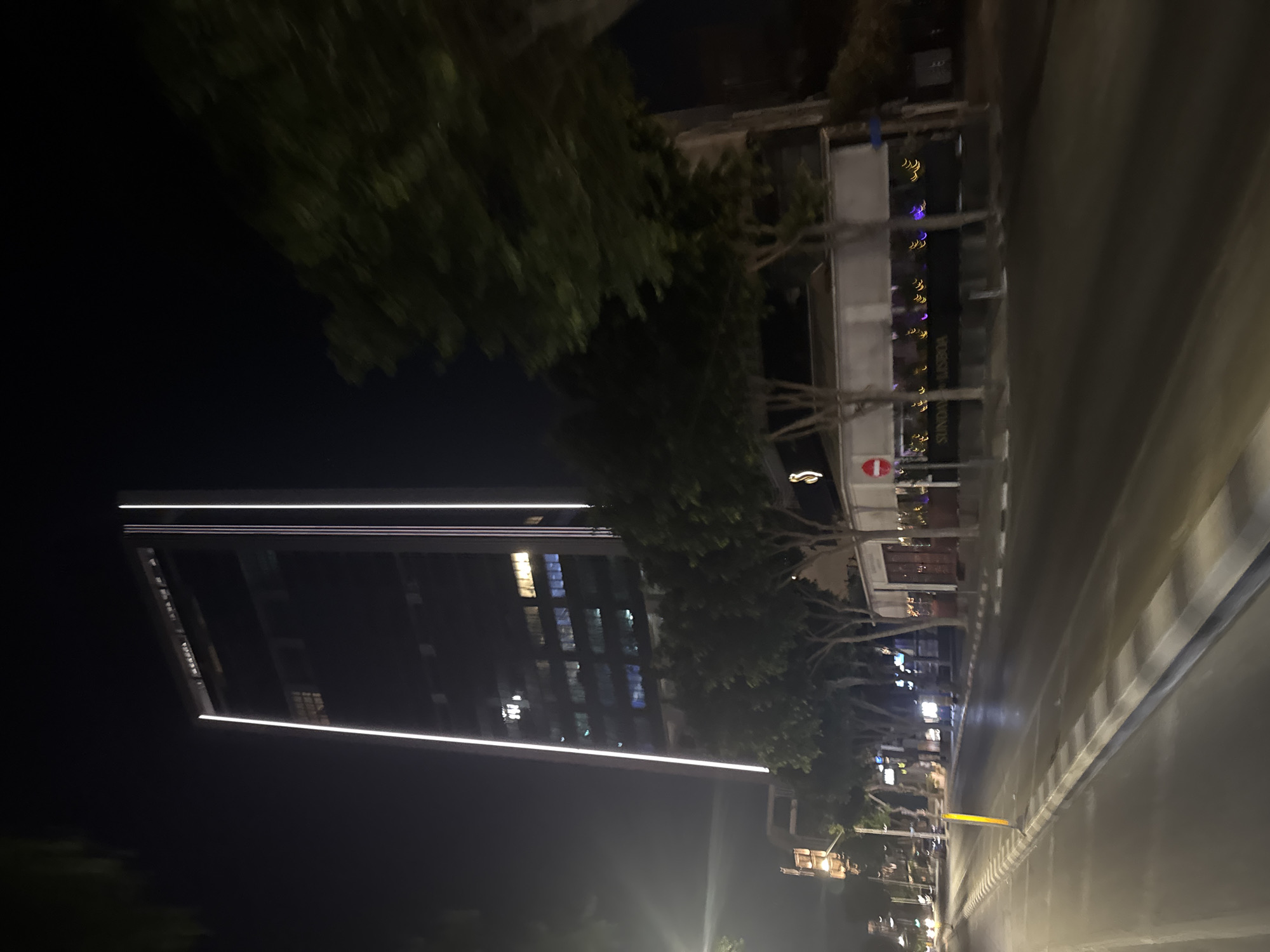 The image shows a street scene at night in Limassol, Cyprus. A modern, tall glass skyscraper dominates the skyline, with vertical white lighting strips running up its sides. In front of the skyscraper, a low-rise building with a dark facade is visible, featuring a sign that reads "SUNDAY LISBOA." Numerous trees line the street, their branches and leaves blurred due to motion. The street itself is wet, reflecting the lights from buildings and vehicles. A yellow traffic cone is present on the side of the road. A traffic light structure is visible in the background to the left. The overall impression is of a lively urban area at night.