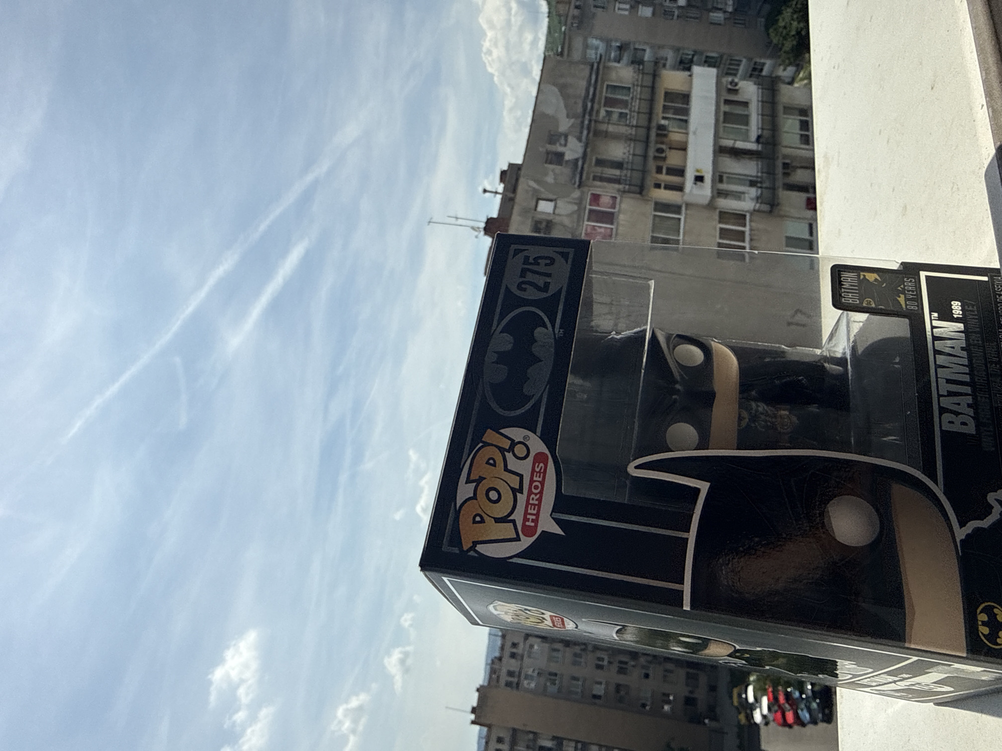 An outdoor, rotated image depicts a Funko Pop! vinyl figure box, likely placed on a balcony or window ledge, set against an urban backdrop. The primary subject is the black box for a Batman Funko Pop! figure, with the character's cowl and eyes prominently displayed on the front.
The scene captures a bright daytime setting with a clear, light blue sky featuring wispy cirrus clouds and distinct white contrails from an aircraft. In the background, several multi-story residential buildings with multiple windows, balconies, and some air conditioning units are visible, suggesting a densely populated urban area. The architecture appears functional and typical of Eastern European cities. A street with parked cars is faintly discernible in the lower background. The foreground shows a plain white wall or ledge, indicating the perspective from an elevated position.
Visible text on the Funko Pop! box includes: "POP!", "HEROES", the number "275", "BATMAN", "TM", "80 YEARS", and "1989". Smaller text also indicates "VINYL FIGURE" and "FIGURINE EN VINYLE". The box features the iconic Batman logo.
The image is captured in Stara Zagora, Bulgaria, evident from the style of the buildings and urban environment.