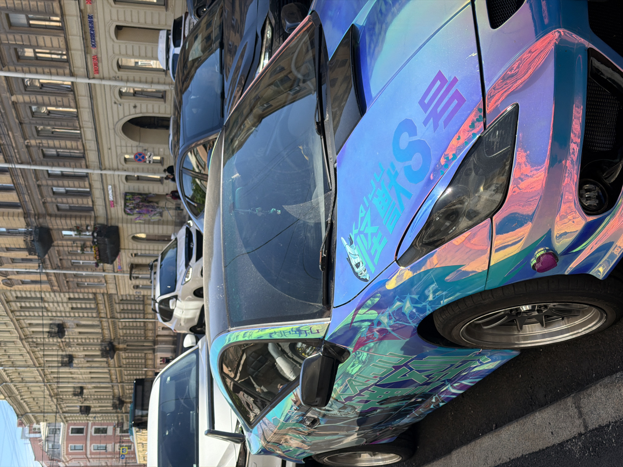 The image shows a street scene in Saint Petersburg, Russia, with several cars parked along a building. The perspective is from a low angle, looking upwards, and the image is oriented sideways.
The primary subject is a highly customized blue car with an iridescent, holographic wrap that shifts colors in the light, displaying shades of blue, purple, and pink. The car has intricate graphic decals, including what appears to be Japanese text, a stylized skull, and abstract patterns. The front headlight and a side mirror are visible, as is a portion of the wheel with a silver rim.
Other vehicles are visible in the background, including a white SUV and a dark-colored car. The buildings lining the street are old European-style architecture with numerous windows and ornate facades. Some of these buildings have signs with text, but the specific words are difficult to discern due to the image quality and orientation. One sign appears to have Cyrillic lettering.
The scene suggests it is daytime, likely sunny, given the bright reflections on the car and the shadows. The atmosphere appears to be casual, with no people prominently featured, though figures can be seen in the reflections and in the background. The overall impression is of a street in a historic European city, with a focus on a uniquely decorated vehicle.