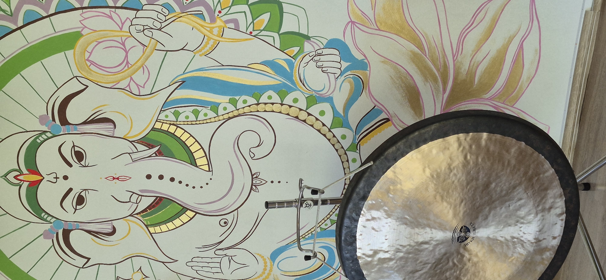 The image shows a painted mural of a deity, likely Ganesha, with a large golden gong positioned in front of it. The mural is depicted in a stylized, illustrative manner with various colors including white, blue, green, gold, and pink. The deity has an elephant head and is adorned with golden jewelry. A lotus flower is also painted on the mural next to the deity. The gong is mounted on a stand and takes up a significant portion of the foreground. The location is Unknown, Unknown.