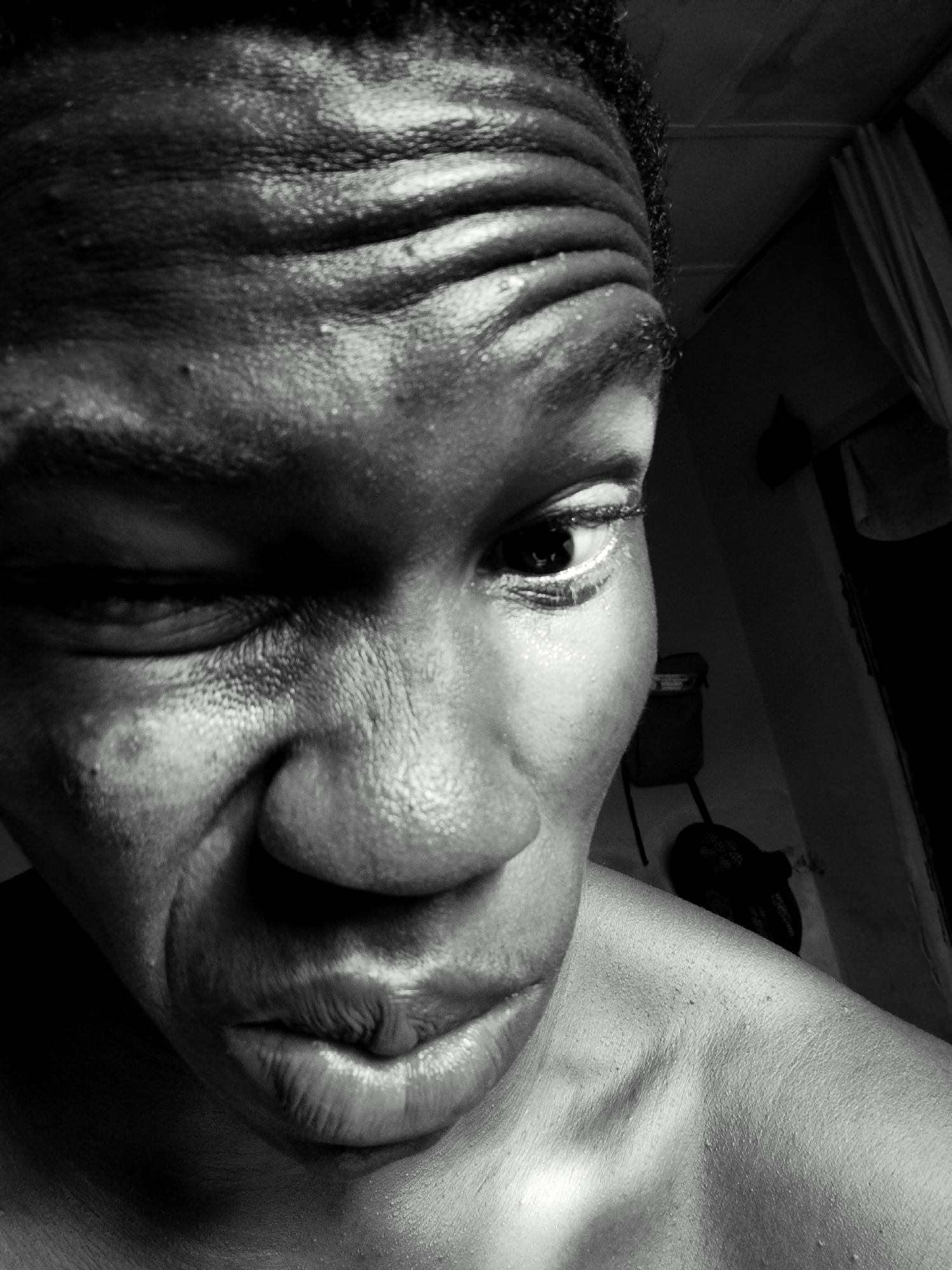The image is a close-up, black and white photograph of a dark-skinned person's face, likely male, angled from above. The person is winking with the left eye almost closed. The forehead has distinct lines and the face appears slightly sweaty. In the background, there is a room with a white wall and some dark objects, including what appears to be a bag hanging on a chair. The location and city are unknown.