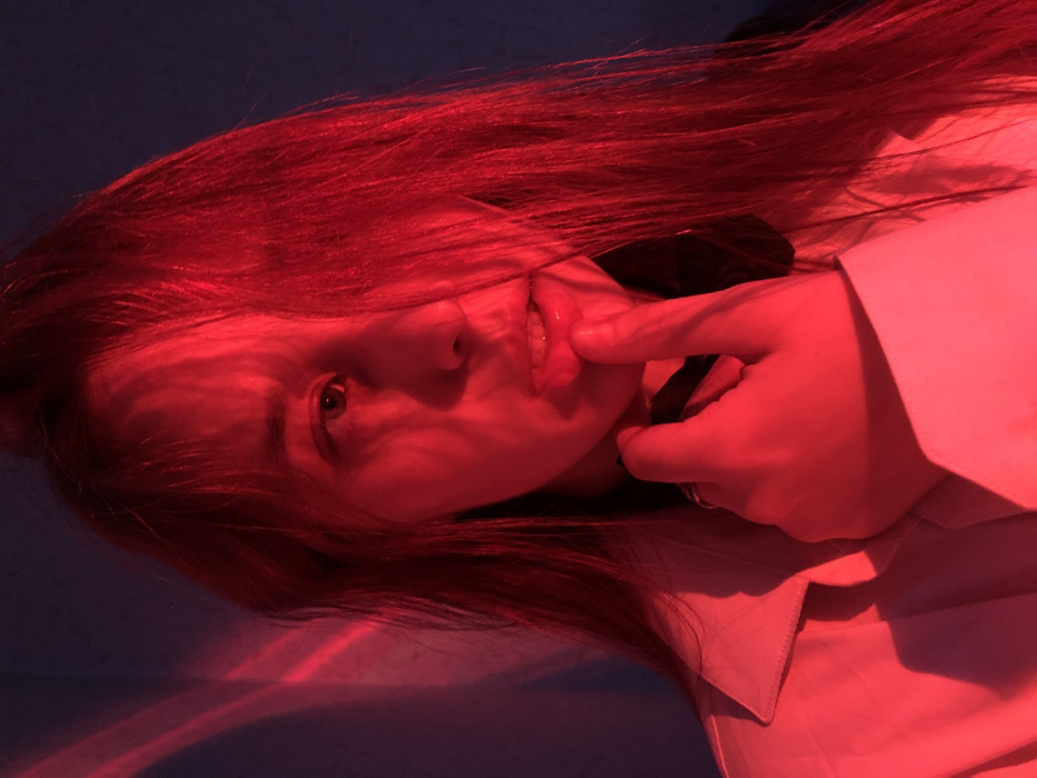 A close-up, chest-up portrait depicts an individual, likely female, illuminated by intense red light emanating from the right, saturating the subject and background in crimson. The subject's long, light brown hair, tinged red, partially covers the left side of their face. Their right eye is visible, directed towards the viewer, while the left side of the face is largely in shadow. The mouth is slightly open, revealing teeth, and the right index finger is pressed against the lower lip. The subject wears a light-colored collared shirt or jacket and a dark band or choker around the neck. The background is a plain, dark-toned wall with distinct, curved red light streaks projected onto its surface. The scene is located in the city of Фоки, Russia.