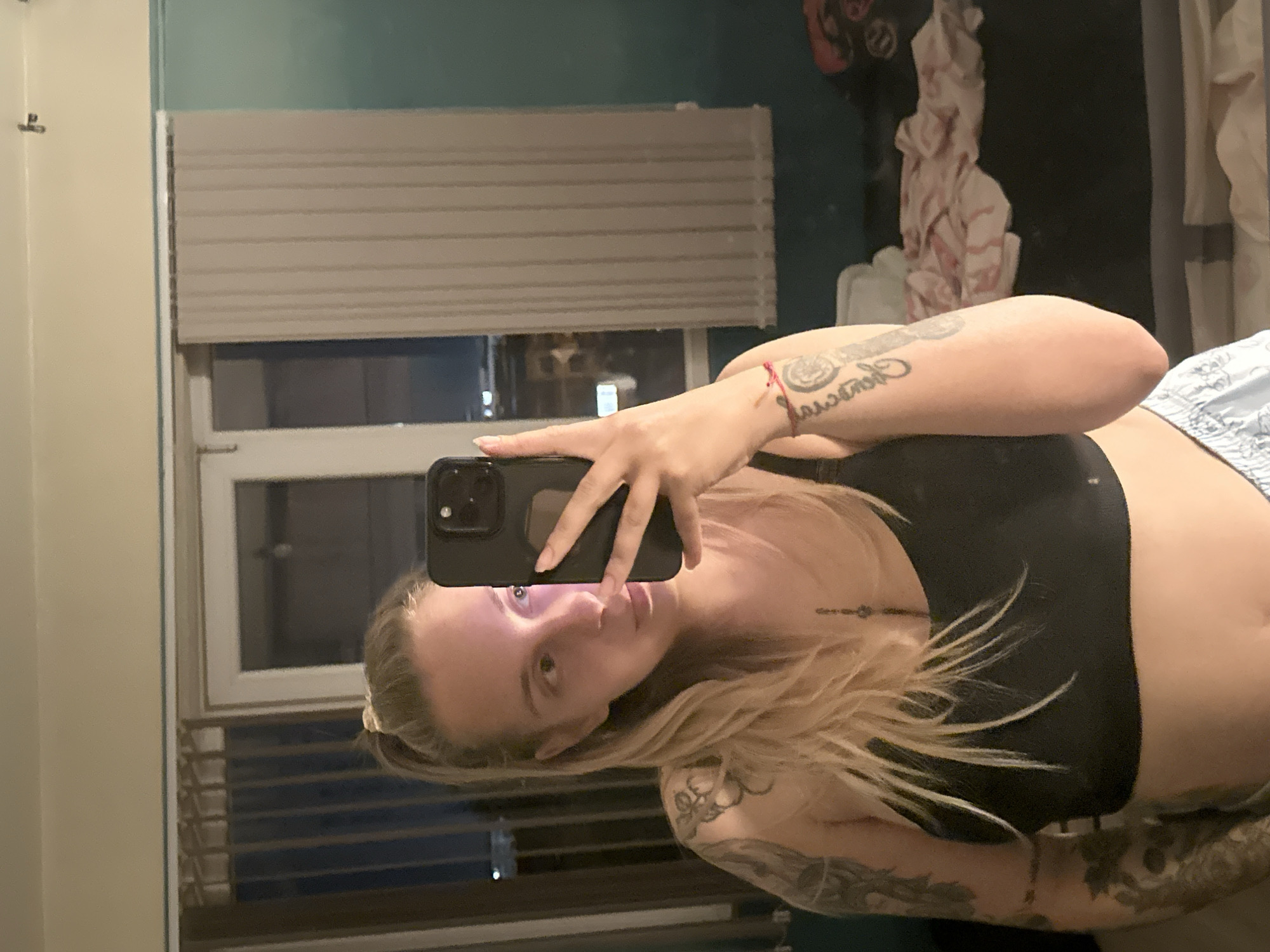 A woman with blonde hair pulled into a ponytail, wearing a black bra and light blue patterned shorts, is taking a mirror selfie in what appears to be a bedroom. She has tattoos on her arms and chest. The room is dimly lit, with vertical blinds covering a window in the background. There is a bed with some fabric on it behind her. The image does not contain any information that allows for its location to be identified as Plovdiv, Bulgaria.