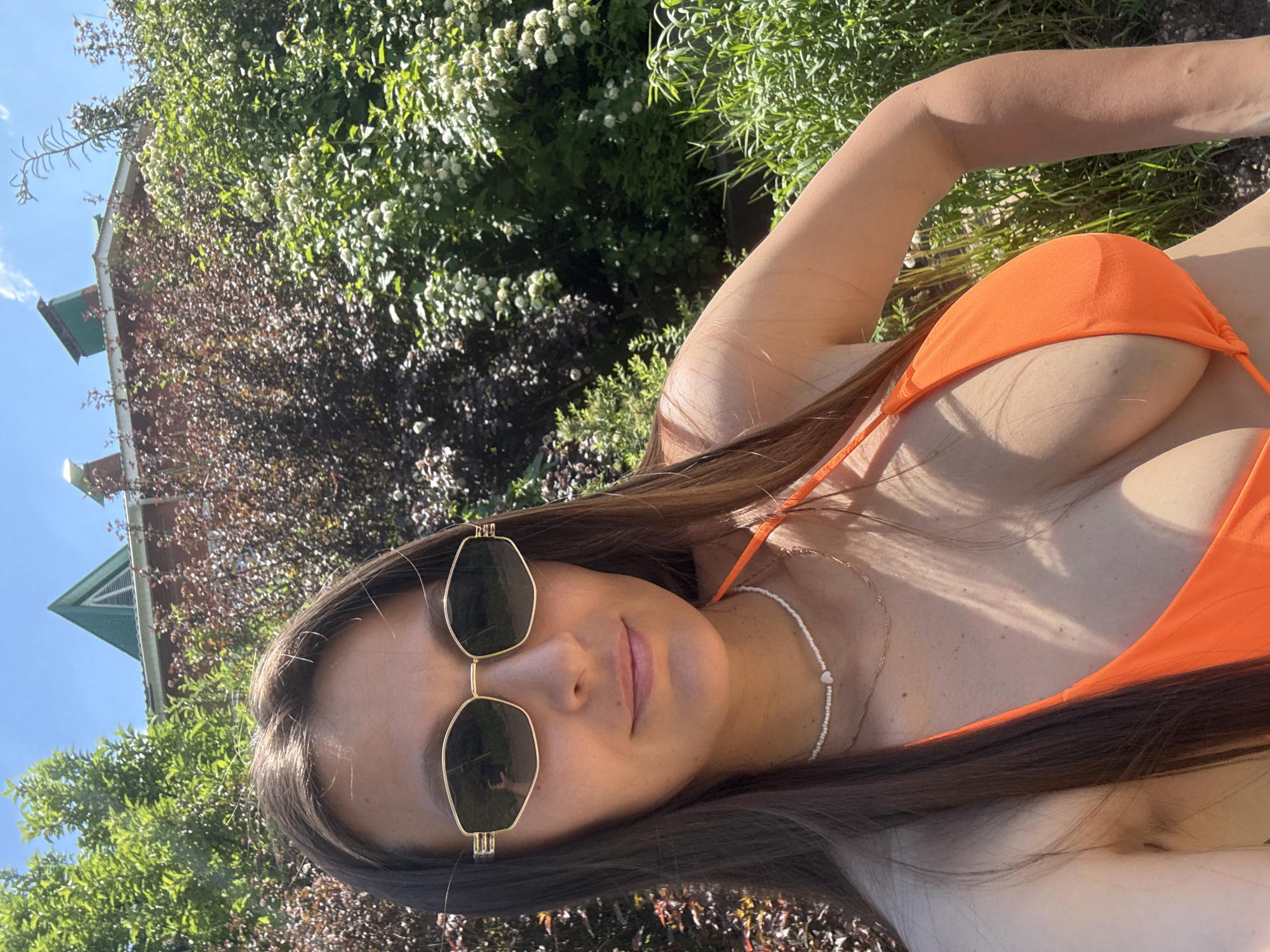 An outdoor, eye-level, close-up, selfie-style photograph captures a young adult female with long, dark brown hair and fair skin wearing an orange bikini top and sunglasses. She is positioned in the foreground, with lush green foliage and a brick building with a green roof visible in the background. The setting appears to be a sunny day, with a clear blue sky overhead. The location is identified as Chaikovsky, Russia.