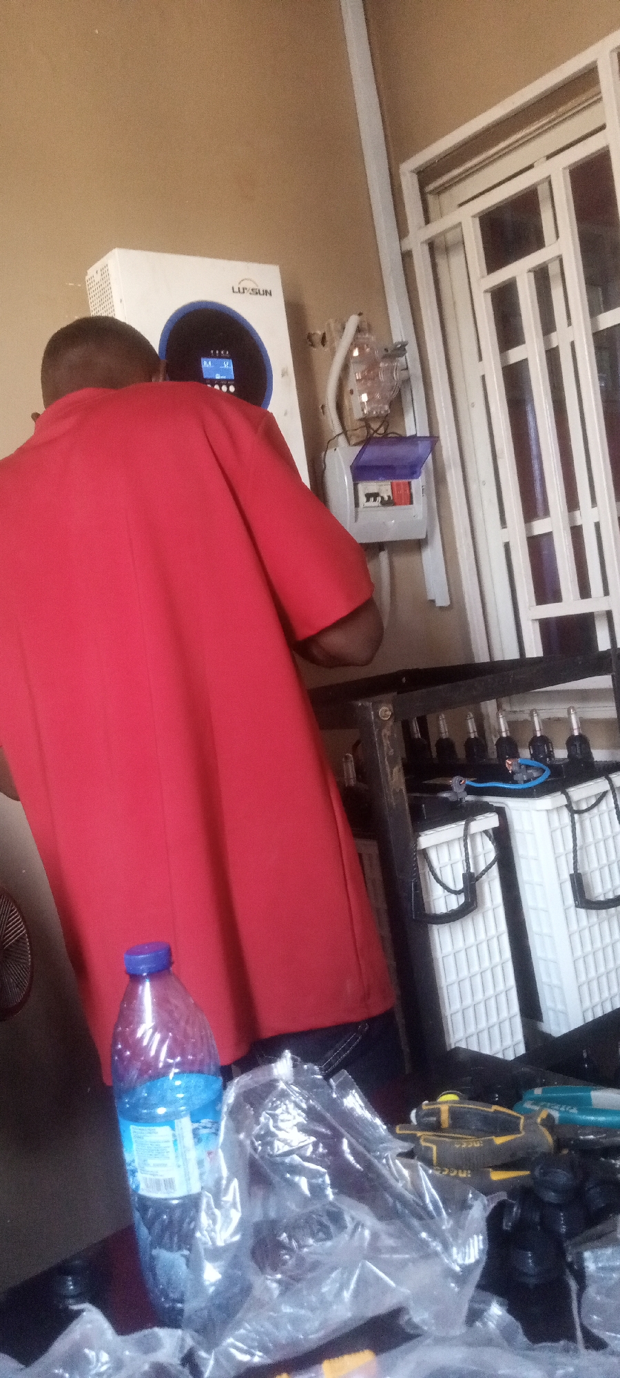 In Sabon Gari Nassarawa, Nigeria, an individual in a red shirt stands near a white electrical box and an inverter. The inverter is labeled "LUXSUN" and has a blue display. Below the electrical box, there is a black metal rack with white batteries. In the foreground, there is a blue water bottle, plastic wrap, and tools on a surface.