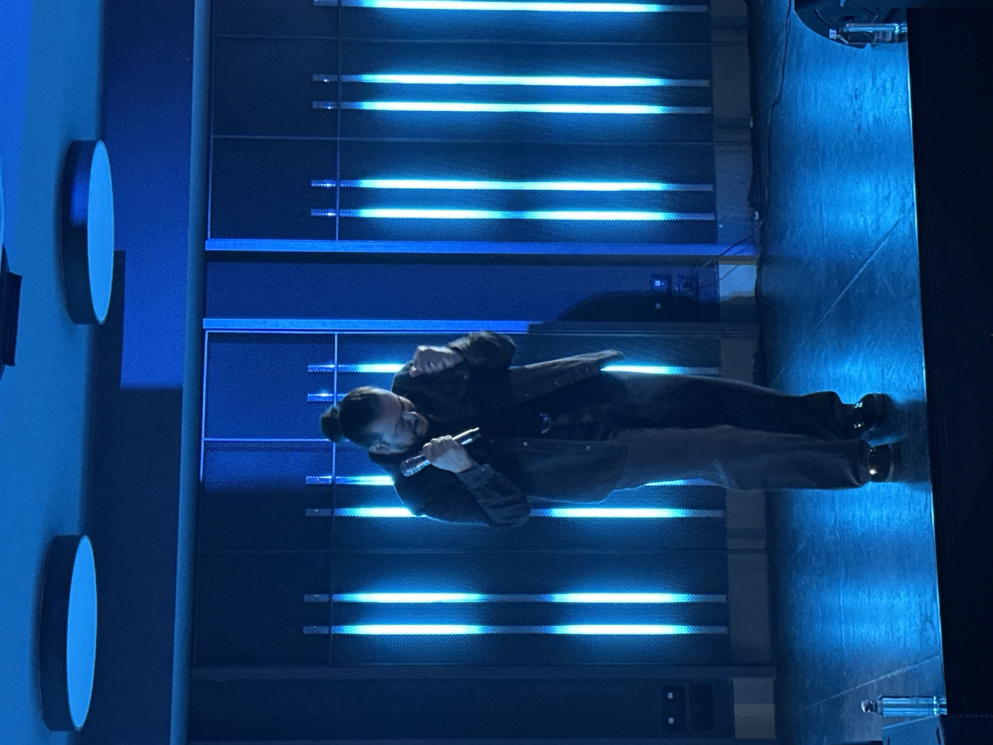 A person with dark hair in a bun, wearing a dark jacket and light-colored pants, is holding a microphone and speaking, possibly singing or rapping, on a stage in Kazanlak, Bulgaria. The stage backdrop features horizontal rows of textured material illuminated by blue light strips. Two circular elements are visible on a vertical surface to the left. The person's feet are visible on a reflective stage surface. The scene is lit with bright blue light.