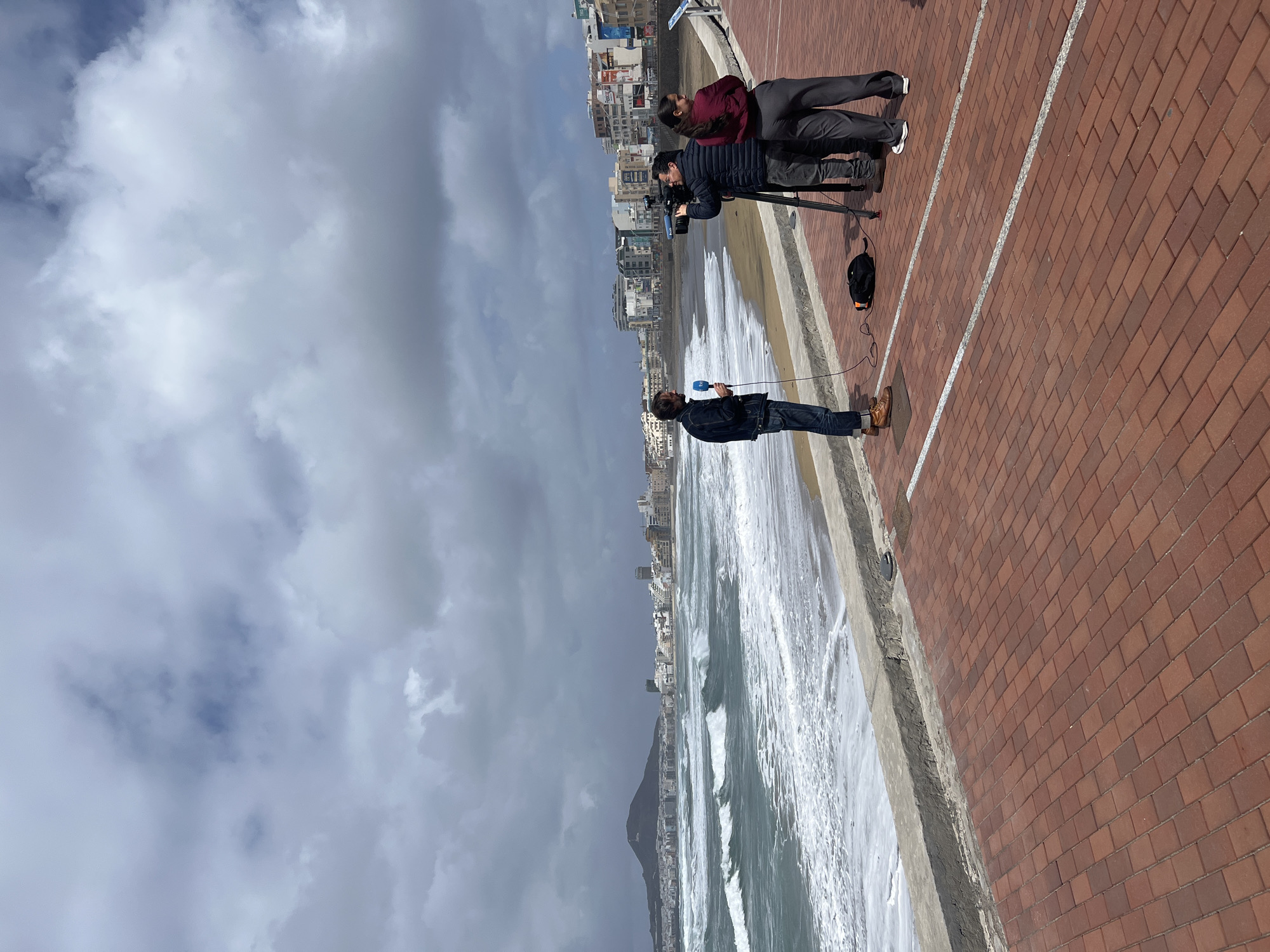 A man is holding a microphone and talking, while a cameraman films him. A woman is standing nearby. They are on a brick promenade overlooking a beach with crashing waves and city buildings in the background. The location is the Playa de Las Canteras in Las Palmas de Gran Canaria, Spain. The sky is overcast.