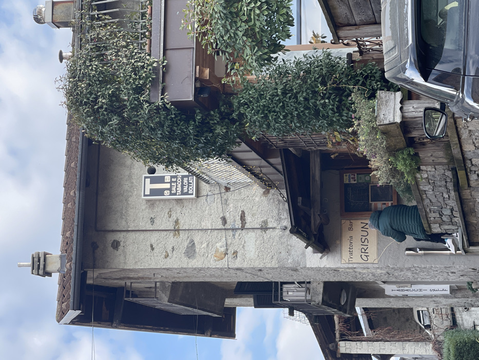 This image depicts a narrow street scene in Castione, Italy, featuring traditional architecture with stone walls, wooden elements, and tiled roofs. The photo is taken during the daytime under a bright sky with some clouds.
On the left, a multi-story building with a brown tiled roof and a chimney-like structure on its peak is visible. Its facade is made of light-colored stone. A prominent black and white sign is affixed to this building, displaying a large "T" and the Italian text "SALE E TABACCHI" (Salt and Tobacco) and "VALORI BOLLATI" (Revenue Stamps), indicating a tobacconist shop. Lush green foliage cascades over a balcony on the upper part of this building.
Towards the right and slightly recessed, another building features wooden balconies and extensive greenery, including vines and potted plants. At street level, a cream-colored sign is mounted on the building, reading "Trattoria Bar" above the name "GRISUN" in larger letters, identifying a restaurant and bar. In front of this establishment, a person is seen from behind, wearing a dark green puffer jacket and dark trousers, bending over near a white pipe or post. The person's face is not visible.
On the far right, the front fender and side mirror of a dark-colored vehicle, possibly a car or SUV, are partially visible, parked along the street. The overall scene suggests a quaint and lively European village setting with commercial establishments.