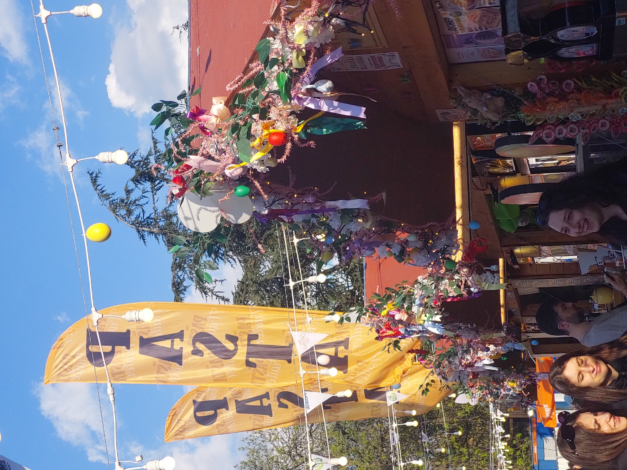 The image shows an outdoor scene in an unidentified city. There are several fair-like elements, including yellow banners with the word "FA2I" printed in bold black letters, strung alongside strands of lights with bare bulbs. These are set against a blue sky partially filled with white clouds.
A red building, possibly a stall or kiosk, is visible, adorned with decorative flower arrangements with ribbons and ornaments. In front of the building, a group of people is present. A woman in the foreground is smiling and looking towards the left of the frame, partially obscured by the banner. Behind her, a man and another woman are also visible near the stall, with only parts of their faces visible. The setting suggests an outdoor event, possibly a fair or festival.