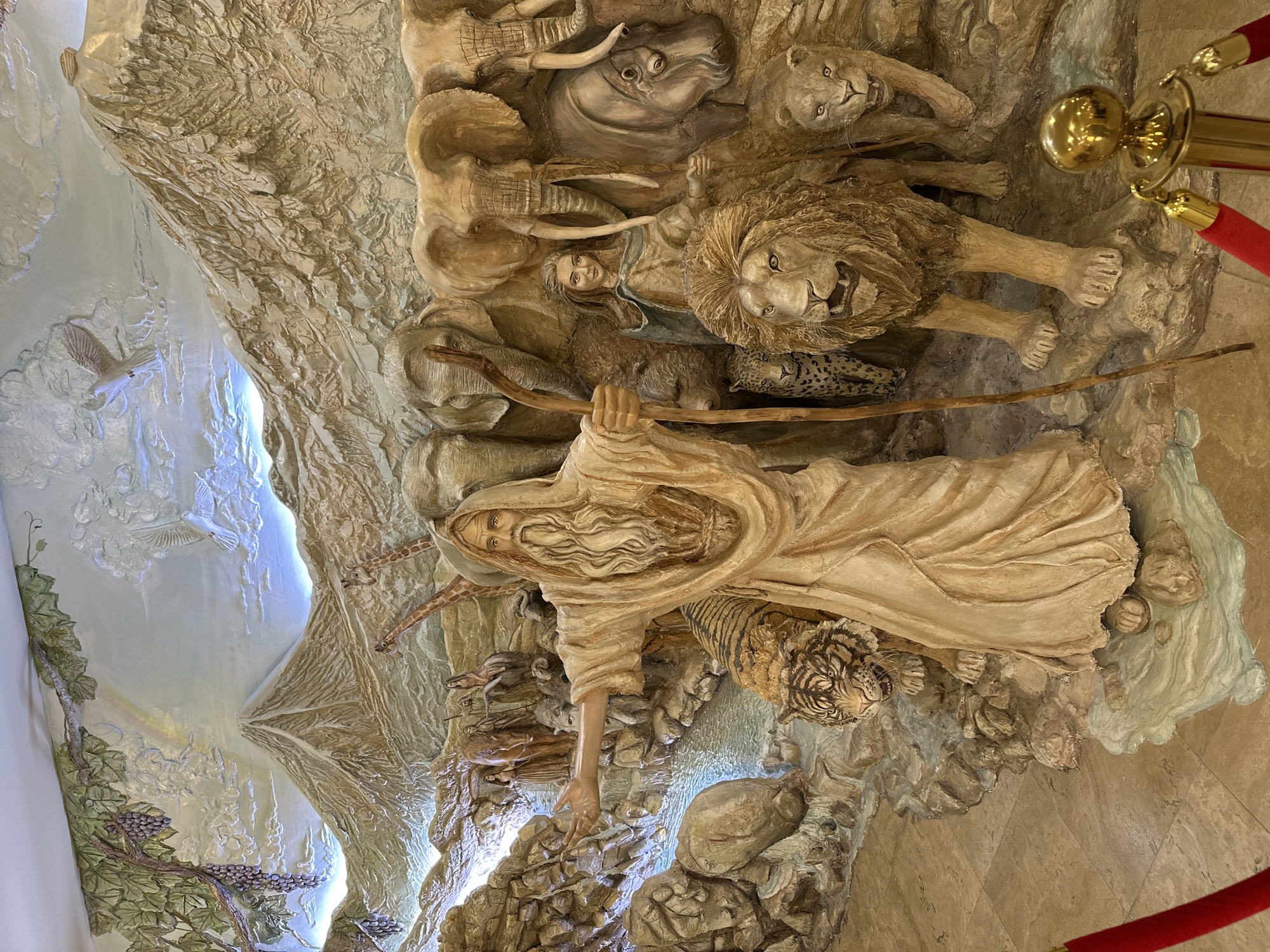 This media file features a large, detailed bas-relief sculpture, likely depicting a biblical scene, possibly Noah's Ark. The central figure is a bearded man, dressed in flowing robes and holding a staff, reminiscent of Moses or Noah. He is surrounded by a diverse array of animals, many appearing in pairs, such as elephants, lions, rhinoceroses, leopards, tigers, giraffes, and deer, along with a flying dove and turtles. Another female figure is visible among the animals.
The sculpture is intricately carved with naturalistic textures for animal fur and skin, and detailed facial expressions. The background of the relief incorporates stylized elements like mountains, a flowing body of water, and vegetation, including grapevines. A subtle rainbow is visible in the upper left, reinforcing the potential theme of Noah's Ark.
The artwork is well-lit by artificial lights, which highlight its three-dimensional depth and contours. The setting appears to be an indoor public space, such as a museum or grand hall, given the presence of a red velvet rope barrier with golden stanchions in the foreground, suggesting it is an exhibit. The sculpture itself is rendered in a light, natural tone, possibly mimicking stone or wood. The image is taken from a slightly elevated perspective, showcasing a significant portion of the monumental artwork. The overall impression is one of grandeur and detailed craftsmanship. No text is visible in the image. The location context is Yerevan, Armenia.