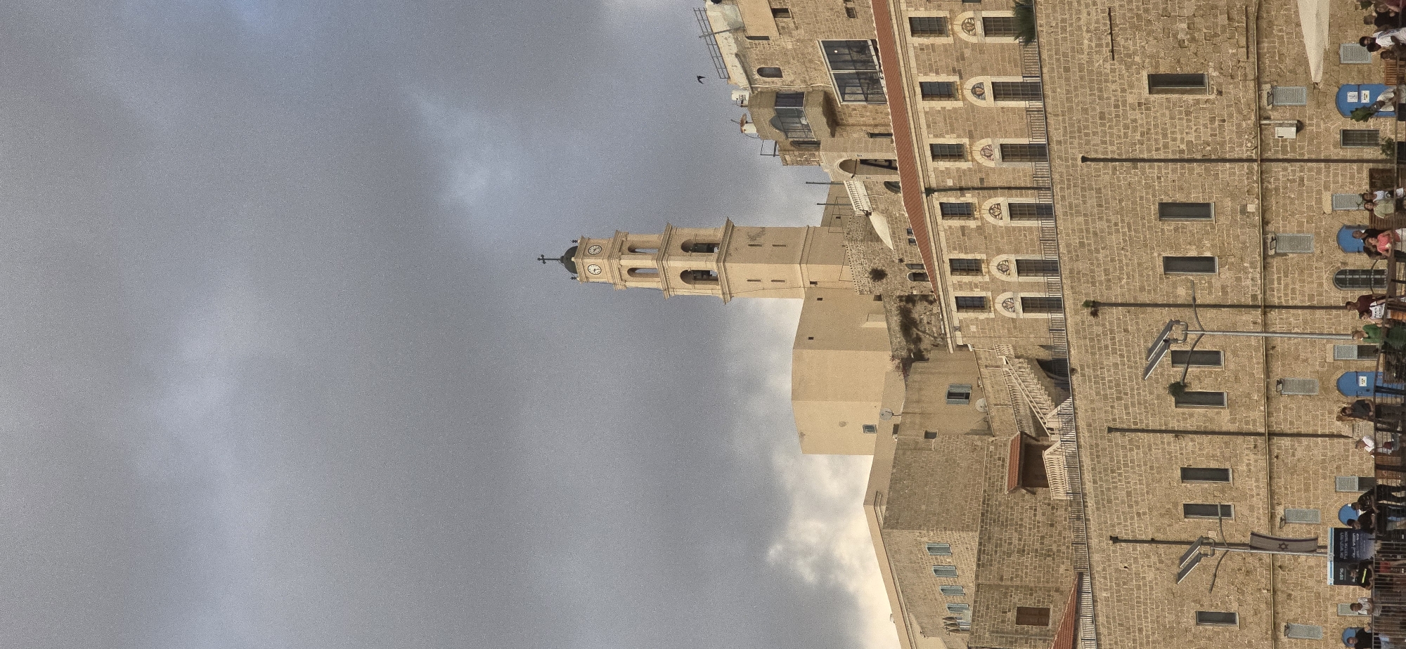 A bell tower, topped with a cross, rises above a cluster of stone buildings under a cloudy sky in Jaffa, Tel Aviv, Israel. The tower has arched openings for bells and clock faces. The buildings are constructed of rough-hewn stone blocks and have red-tiled roofs. A stone staircase leads up the side of one building. In the foreground, a railing separates a street level from the buildings. People are visible on the street below, some gathered near blue doorways. Streetlights and a flag with a Star of David are also present. The sky is dark and overcast, suggesting impending rain or a storm.