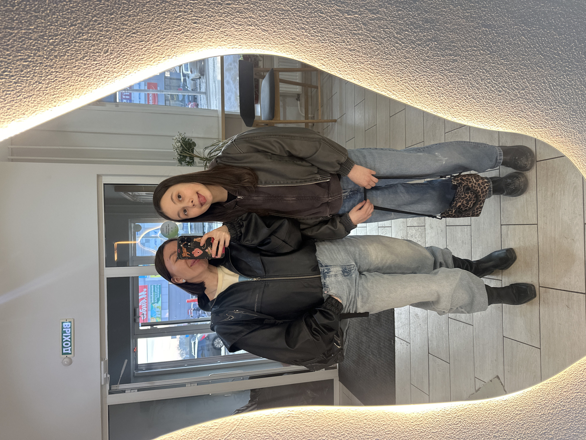 Two females are captured in a full-length mirror selfie taken indoors, reflecting an outdoor street scene. The image displays the interior of a commercial or public establishment in Votkinsk, Russia.
The individual on the left, with dark hair, is wearing a black hooded jacket over a light top, light blue baggy jeans, and black ankle boots. She holds a smartphone with a multi-patterned case in her right hand, taking the photograph. She is smiling with her head tilted. A black bag is slung over her left shoulder.
The individual on the right, also with dark hair, is wearing a dark gray bomber jacket over a dark top, light blue baggy jeans, and dark boots. She holds a small leopard-print handbag in her left hand and is sticking her tongue out playfully.
The immediate indoor environment features a light-colored tiled floor, a high-top table, and a black-seated wooden bar stool. A green potted plant with white flowers is visible behind the right individual.
Beyond a glass door/window, an outdoor street scene is visible. Parked vehicles, including a dark SUV and a white car, are present on what appears to be a cold or snowy street. Reflecting in the mirror above the door is an illuminated green sign displaying the Cyrillic word "ДОХІД" (DOKHID). Further outdoor reflections reveal a partial sign with Cyrillic text, including "Воткинск," confirming the city.