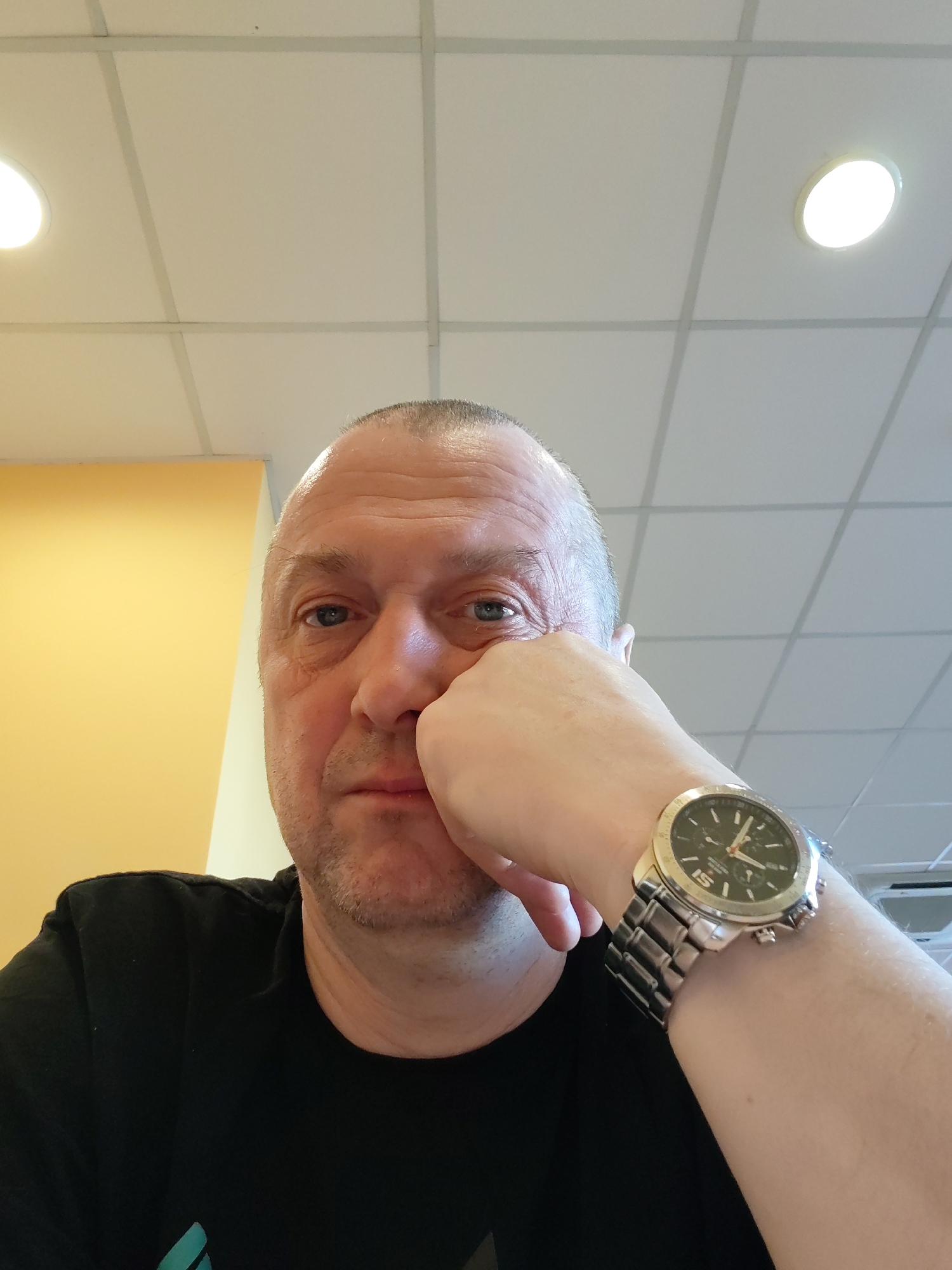 A man is captured in a close-up, eye-level selfie, looking directly at the camera with a neutral to contemplative expression. He is a middle-aged individual with short, light hair, blue eyes, and light stubble. He wears a black t-shirt. His right hand is raised, with his fingers resting against his cheek and chin, appearing to support his head. On his right wrist, he wears a silver-toned wristwatch with a dark circular dial, silver-colored hour markers (including a prominent "5"), and three small sub-dials, indicating a chronograph. The watch has a matching metal bracelet.
The setting appears to be an indoor space with artificial overhead lighting. The ceiling consists of white acoustic tiles arranged in a grid pattern with grey supports. Two bright, circular recessed light fixtures are visible in the upper part of the frame. To the left, a portion of a light yellow wall or partition is visible, suggesting a divided space. No specific activity or event beyond posing for the photo is discernible. The lighting is bright and even, consistent with an indoor environment during any time of day. No explicit text other than the numbers on the watch face is clearly readable.
