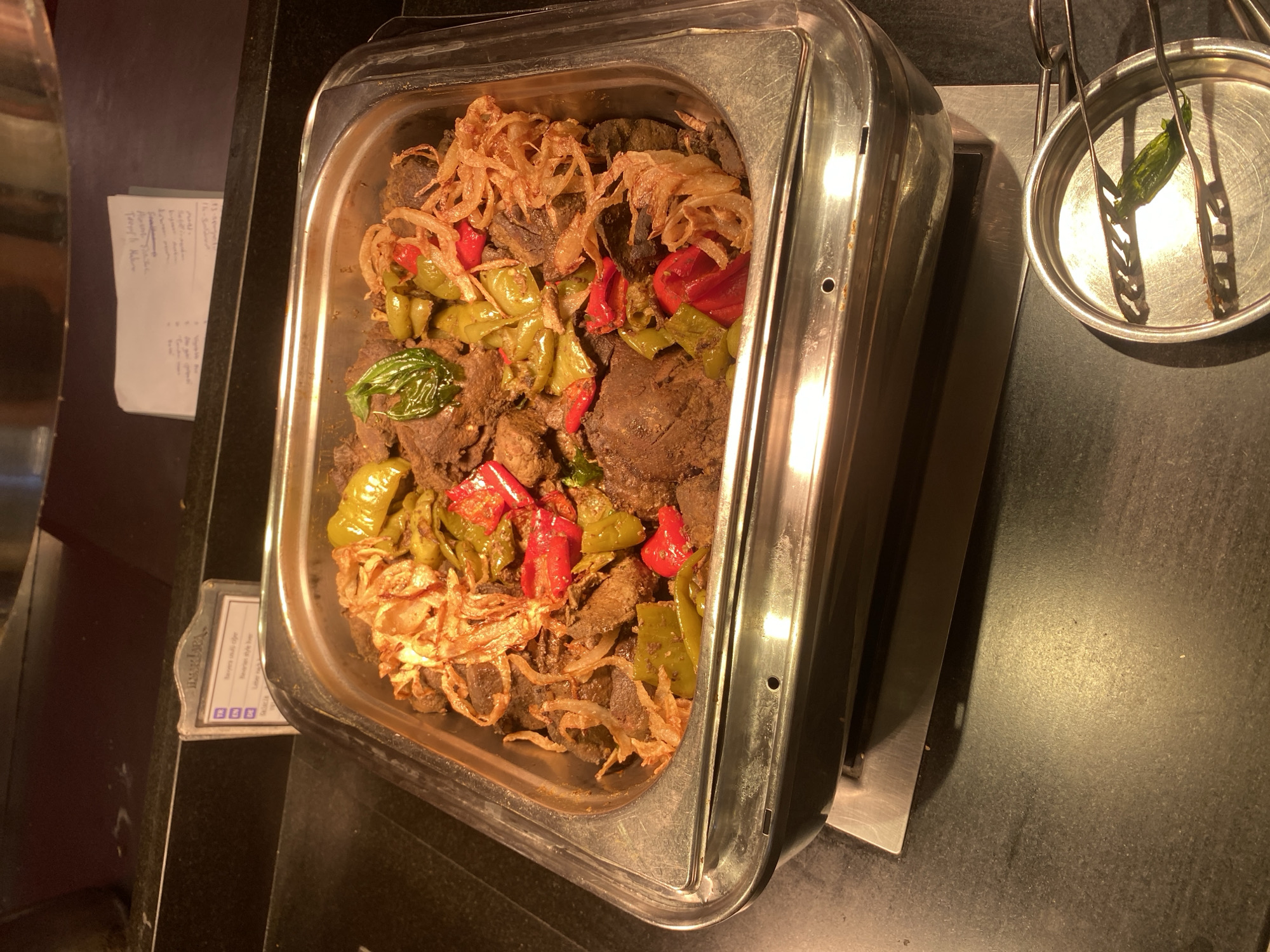 In Aksu, Turkey, a chafing dish filled with a beef stew is displayed on a dark countertop. The stew contains chunks of dark meat, sliced green and red bell peppers, and fried onions. A small metal sign with writing in Turkish and English is placed next to the dish. In the foreground, a small metal dish holds a single green pepper, held by tongs. The scene appears to be in a restaurant or buffet setting.