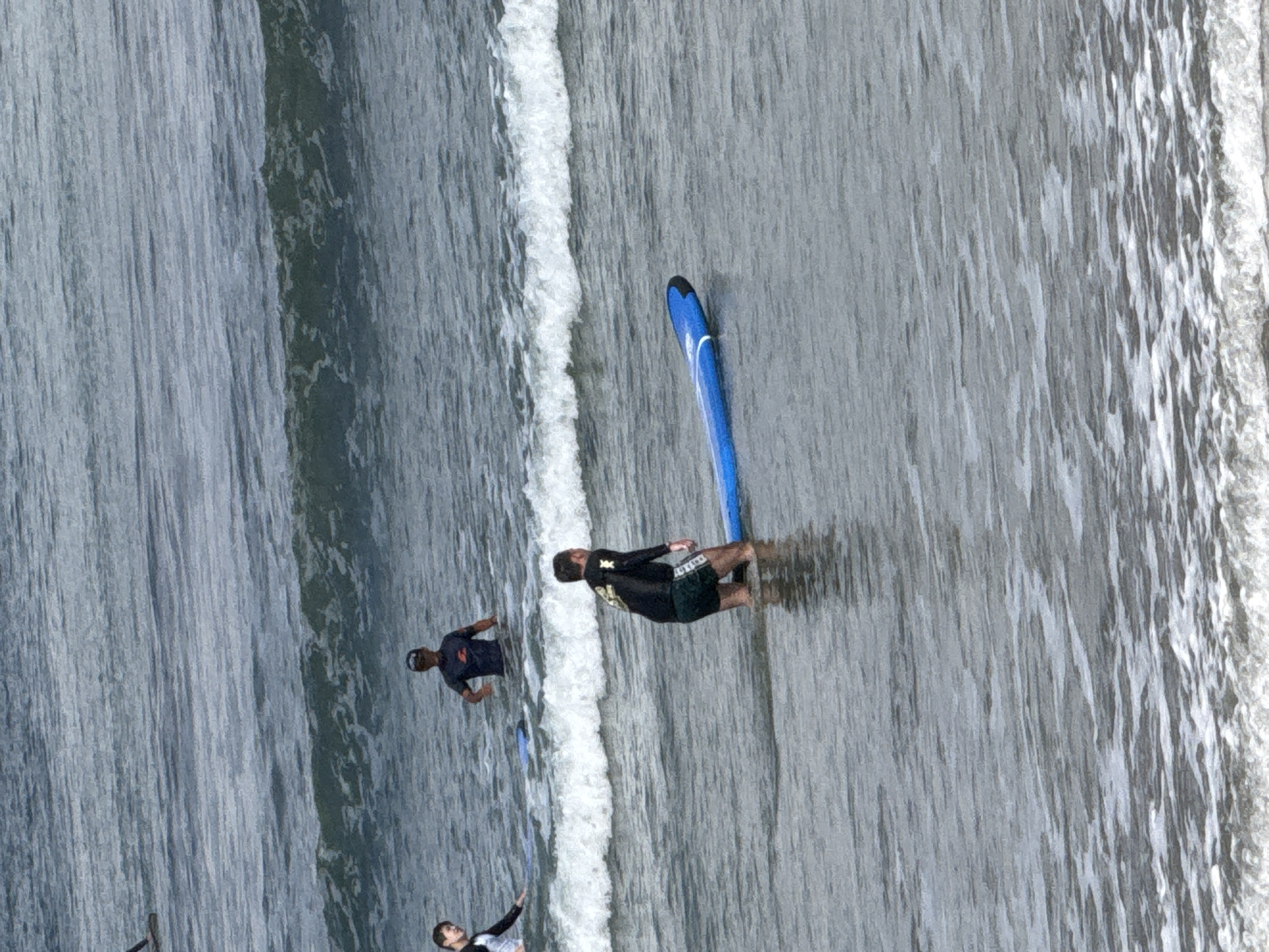 In Kuta, Indonesia, three surfers are in the ocean. One surfer, wearing a dark long-sleeved rashguard and green shorts, kneels in the water with a blue surfboard in front of him. Another surfer, wearing a dark t-shirt and a cap, stands in the water further back, facing the camera. A third surfer, wearing a white and black rashguard, is visible on the left side of the frame, also in the water with a surfboard. The ocean has moderate waves and a white wash breaking on the shore.