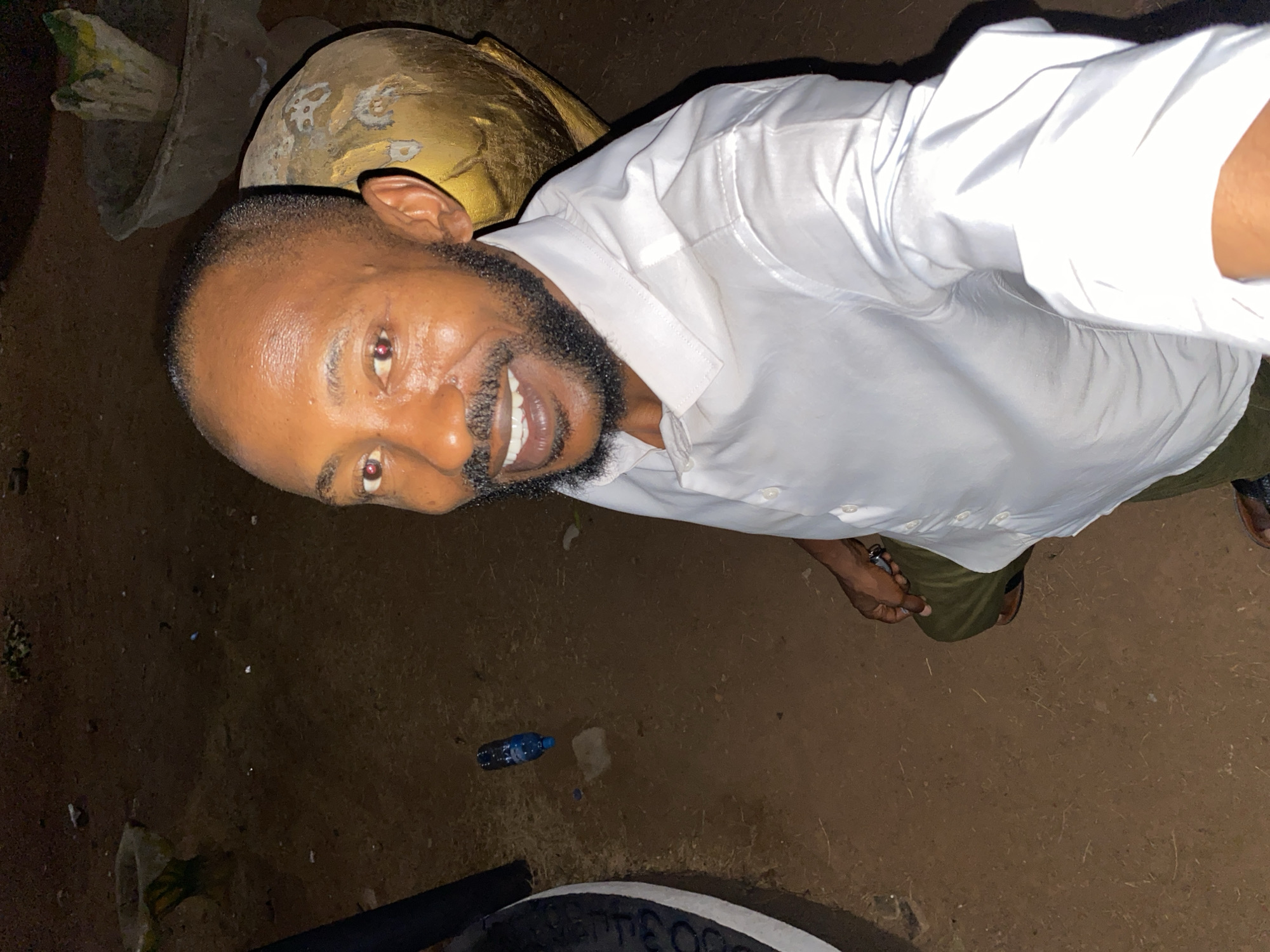 A man with a beard and wearing a white shirt is the subject of a selfie taken in Sabon Tasha, Nigeria. He stands on reddish-brown dirt near a discarded blue water bottle and a black tire. He smiles into the camera. He has something gold by his head.