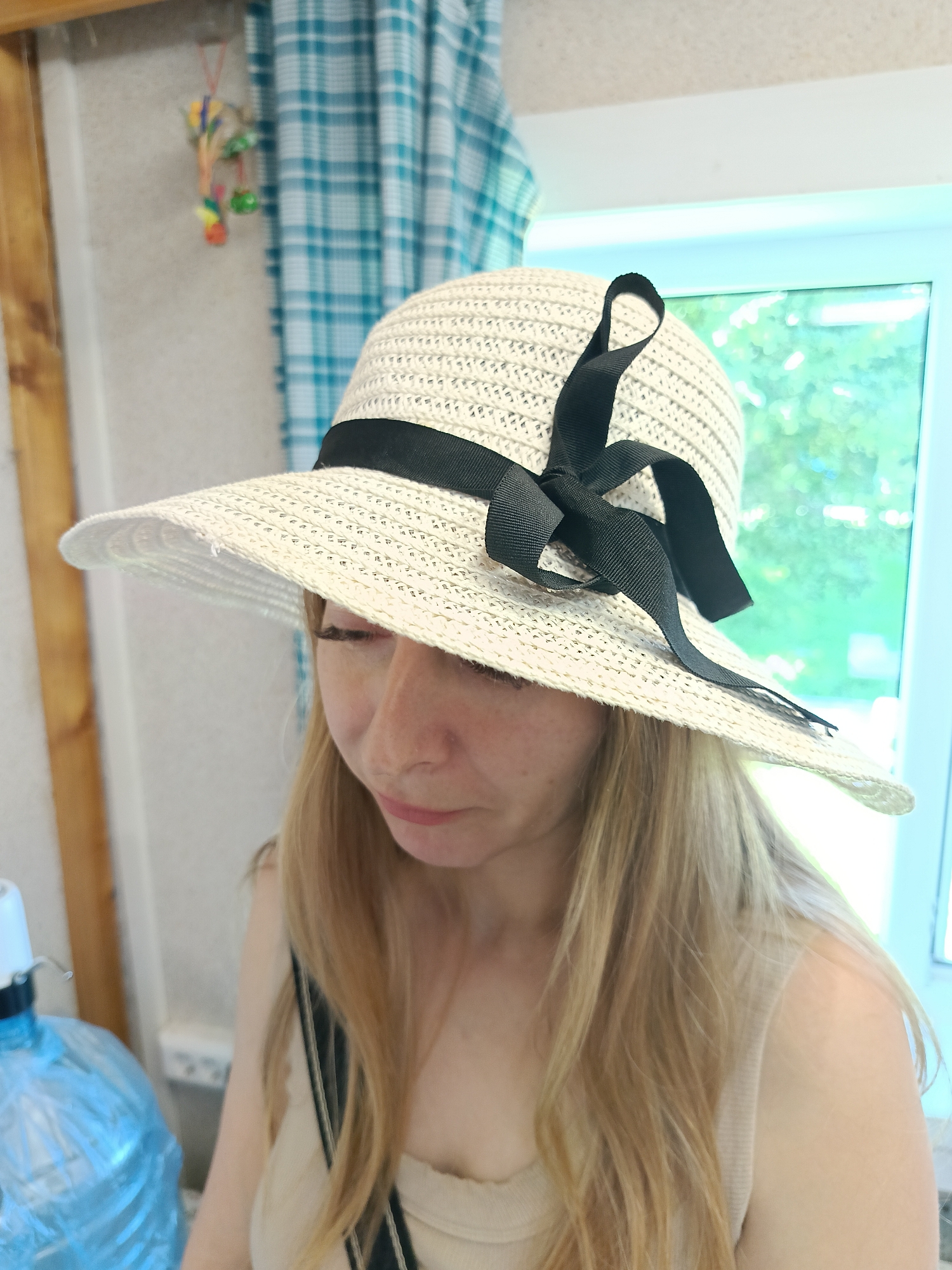 A woman is wearing a straw hat with a black ribbon. She is looking down and her hair is blonde. The background is blurred and shows a window with trees outside, a blue and white checkered curtain, and some decorations hanging on the wall. A blue water bottle with a dispenser is visible on the left side of the image. The scene appears to be indoors. The location is specified as Semenkovo, Russia.