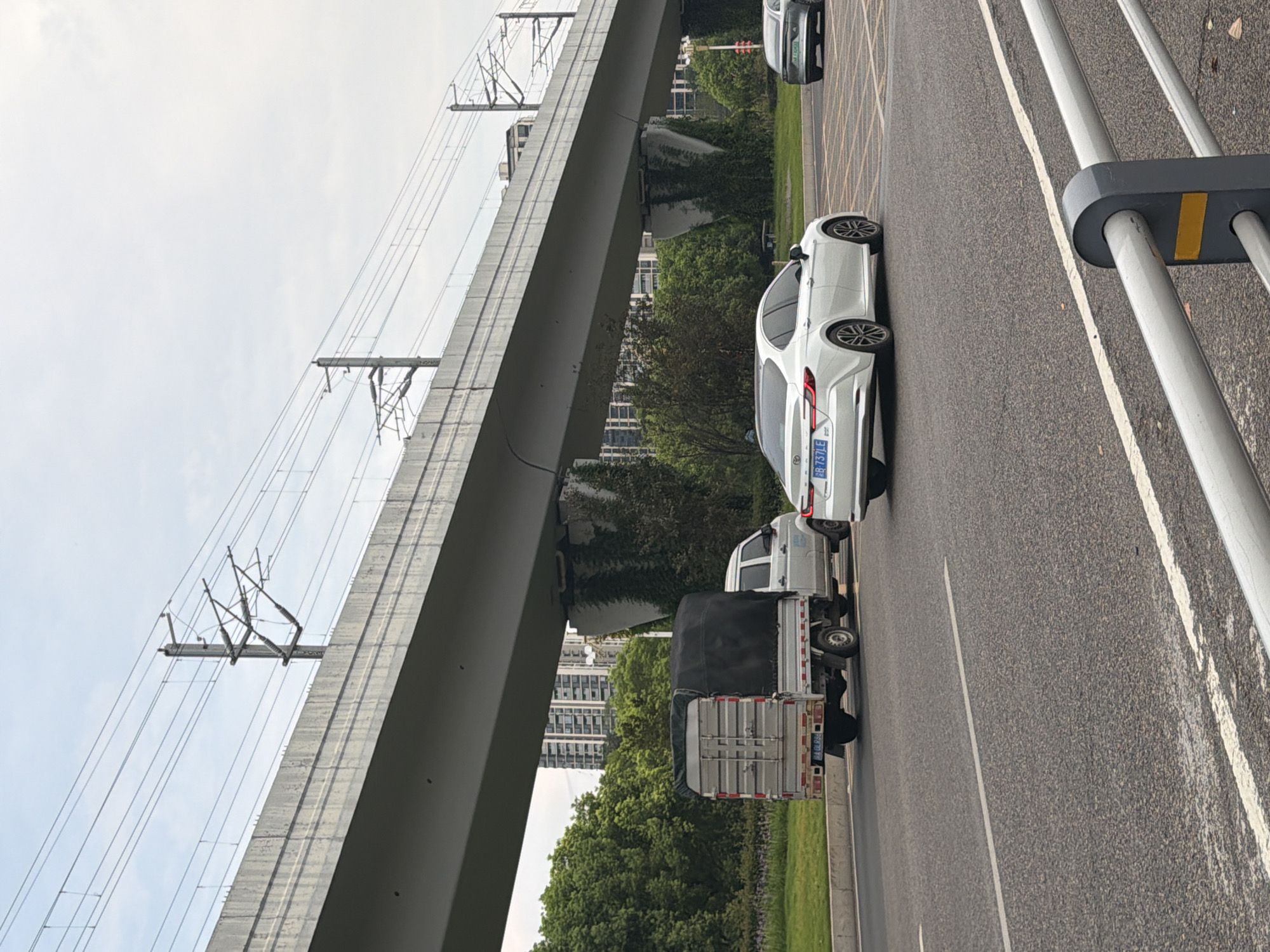 An overpass for a high-speed rail line stretches across a cloudy sky, supported by concrete pillars overgrown with vines. Below the overpass, a gray, tarp-covered truck with a silver cargo box is driving on an asphalt road. To the right of the truck, a white sedan is also traveling on the road. Further in the background, a gray sedan is visible. Trees and tall buildings line the side of the road. This scene is located in the city of Jiangjin, China.