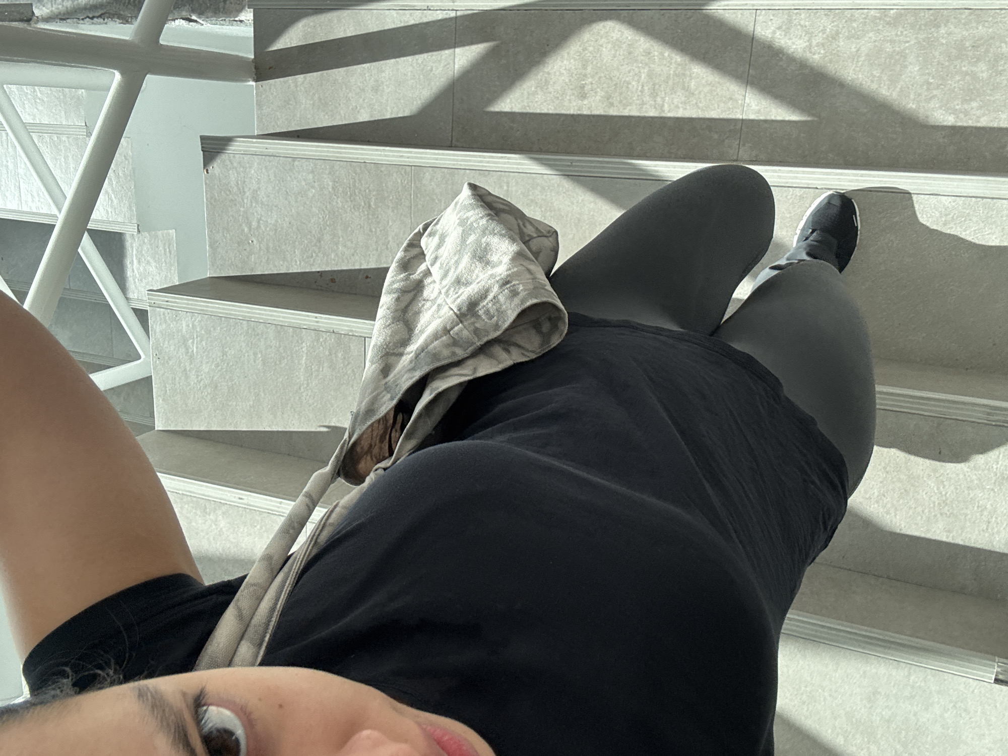 An overhead, high-angle selfie captures a person standing on a light grey staircase in what appears to be an indoor or covered outdoor architectural space. The individual, seen from the chest down, is wearing a black top or t-shirt and dark-colored leggings or shorts. One black sneaker is visible on their right foot, which is placed on a step. A light-colored, patterned fabric item, possibly a tote bag or a folded garment, is draped over their left side. In the bottom left corner, a portion of the person's face, including an eye, eyebrow, and lips, is visible, suggesting they are holding the camera.
To the left of the image, a white metal railing with a distinct geometric or criss-cross design runs alongside the stairs. The stairs themselves are constructed from a light grey material, possibly textured stone or large tiles, with clear, defined edges. Strong, sharp shadows cast by the sun diagonally across the steps indicate bright daylight conditions. The overall scene conveys a modern and minimalist aesthetic. No text is visible in the image. The setting is consistent with a location in Puebla, Mexico.