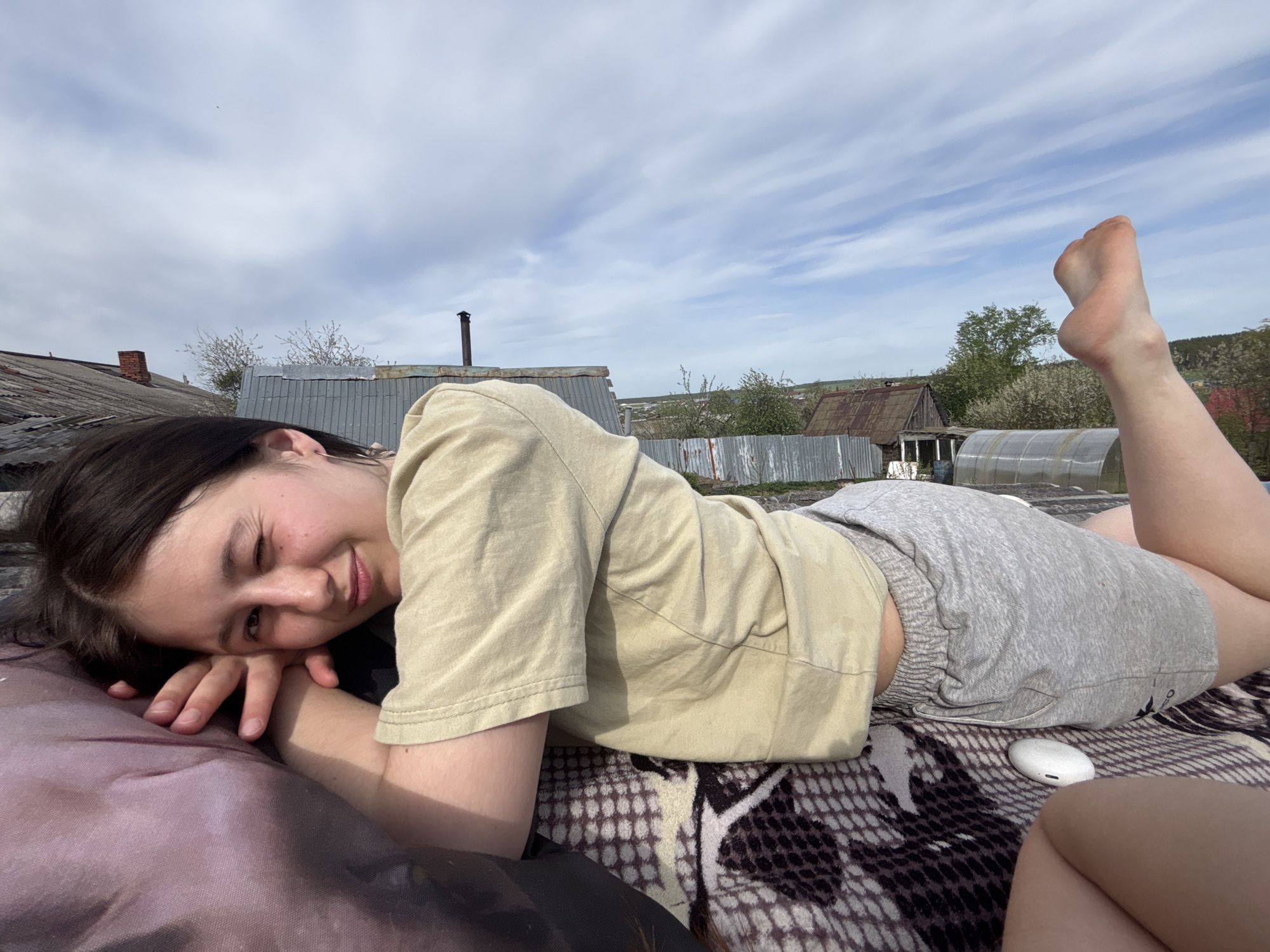 A young woman is lying on a patterned blanket outdoors. She is positioned at an angle in the frame, with her head resting on her hands and her legs extended. She is wearing a light brown t-shirt and gray shorts. Her right leg is raised in the air, exposing her foot. The woman is smiling and looking towards the camera.
Behind her, there is a landscape featuring buildings and a cloudy sky. There is a visible chimney and various structures indicating a rural or suburban environment. A greenhouse-like structure is present in the background.
The presence of what appears to be a pair of wireless earbuds suggests a modern element in contrast with the rustic setting.
Based on the prompt, the location is Фоки, Russia.