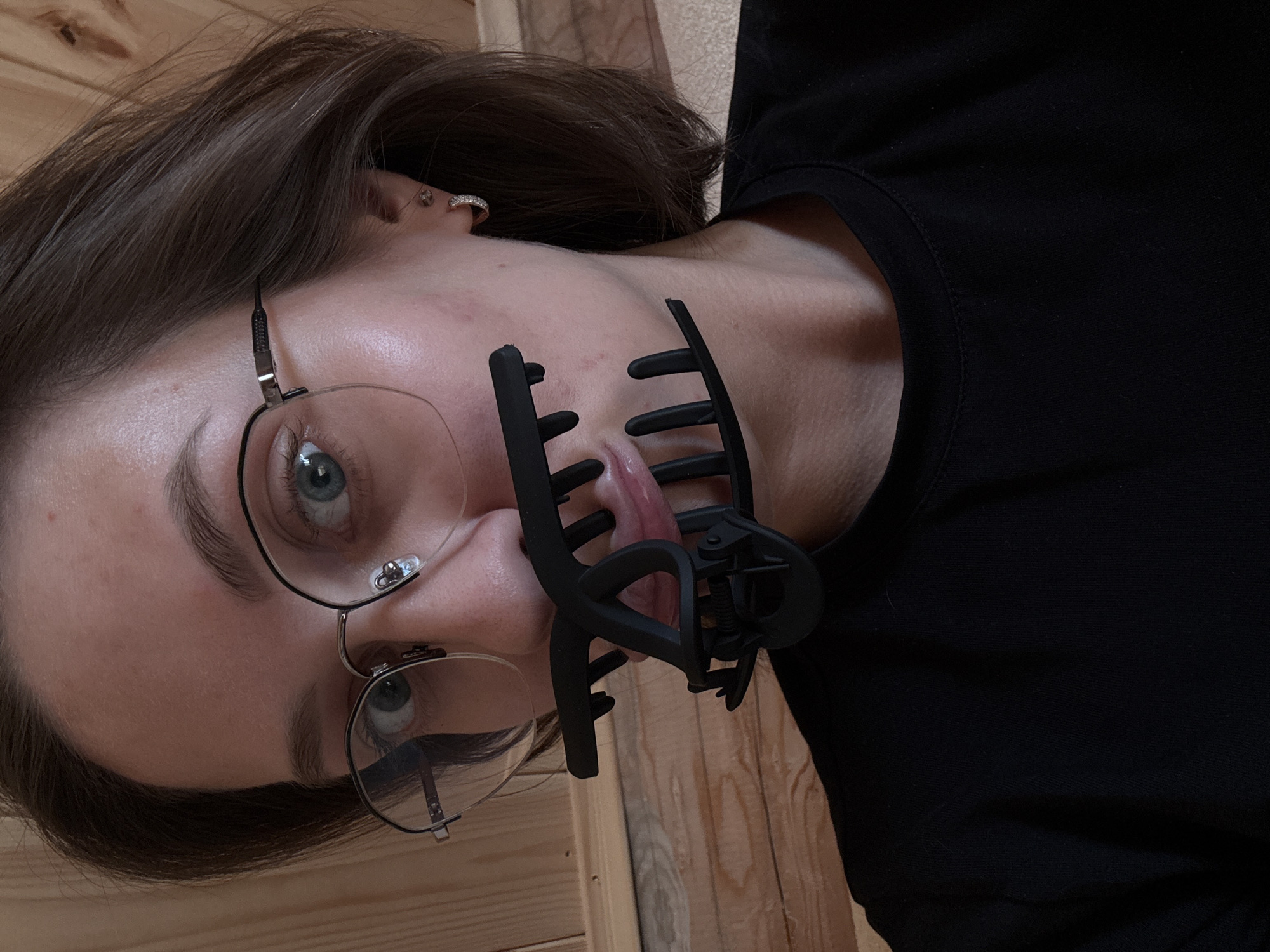 The image depicts a female individual in the city of Lenino, Russia, captured from the mid-chest upwards. She has medium brown hair and wears round-framed eyeglasses. Her light-colored eyes are directed towards the viewer. She exhibits fair skin with visible pores and slight facial redness. Her ears each feature a small hoop earring, with an additional stud in her right ear. She wears a black crew-neck garment. A large, black, plastic hair claw clip is positioned in her mouth, with her tongue protruding and holding the clip's "teeth" apart. The background consists of vertical wooden paneling on the left and a lighter, textured surface on the right.