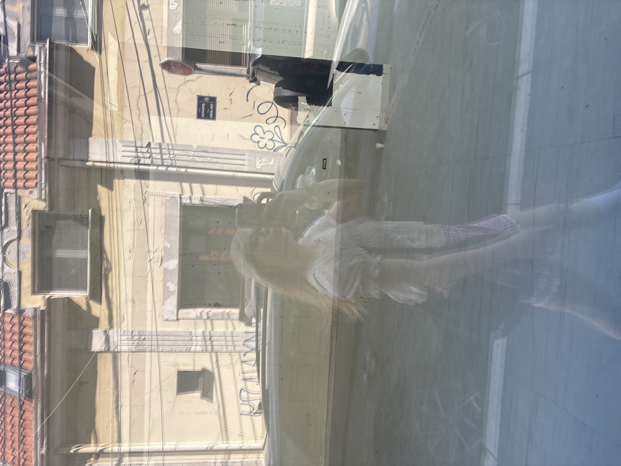 This media file displays a reflection on a glass surface, showing an outdoor urban scene. The main subjects are a woman with blonde hair wearing a white top, and what appears to be a reflection of a car. The scene depicts a building with a tiled roof and cream-colored walls, featuring windows and some signs. A stop sign is visible on the wall, along with a street sign that reads "KRALJA MILUTINA". There is also a drawing of a flower on the wall. The overall impression is of a sunny day in a European city, possibly Belgrade, given the street name and location context. The reflection is somewhat distorted, adding to the surreal quality of the image. The time of day appears to be daytime, with clear lighting.