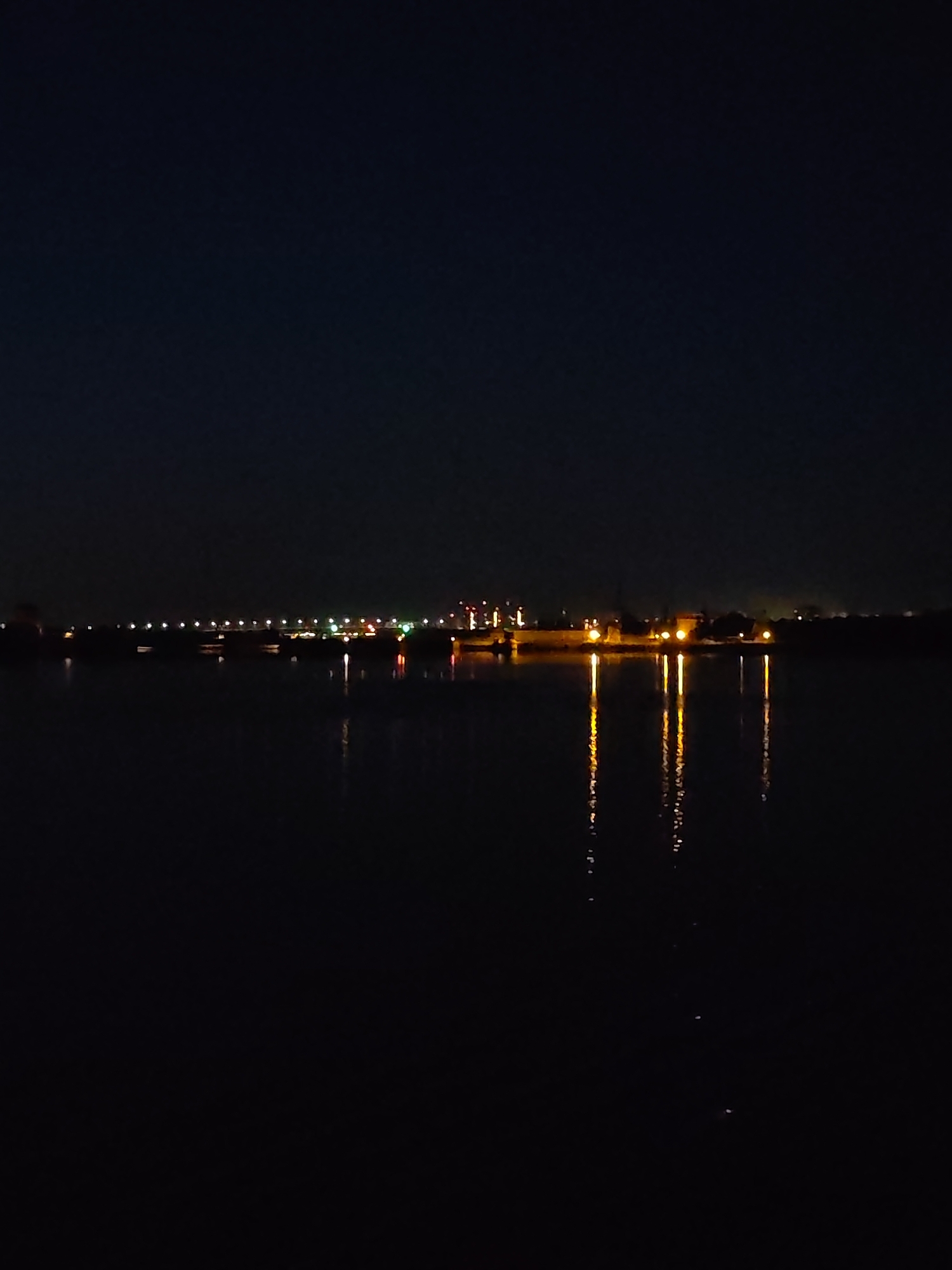 The image depicts a nighttime scene in Dubna, Russia, focused on a wide expanse of dark, calm water, likely a river or canal. The sky above is a deep, unadorned black, devoid of celestial bodies.
Across the midground, a long, horizontal structure, consistent with a bridge or dam, is visible, marked by a series of evenly spaced, small, bright white and yellow lights. These lights cast subtle, diffuse reflections on the water directly beneath them.
To the right of the frame, and appearing slightly closer than the aforementioned structure, is a distinct cluster of intensely bright, orange-yellow lights originating from a landmass or prominent structure on the far bank. These lights produce vivid, elongated vertical reflections that shimmer brightly on the dark water's surface. Interspersed with these bright clusters and extending further into the distance are additional scattered pinpoints of light, some green and red, indicative of an illuminated cityscape or industrial area along the distant bank. The immediate foreground consists solely of the dark water. No people or specific actions are discernible.