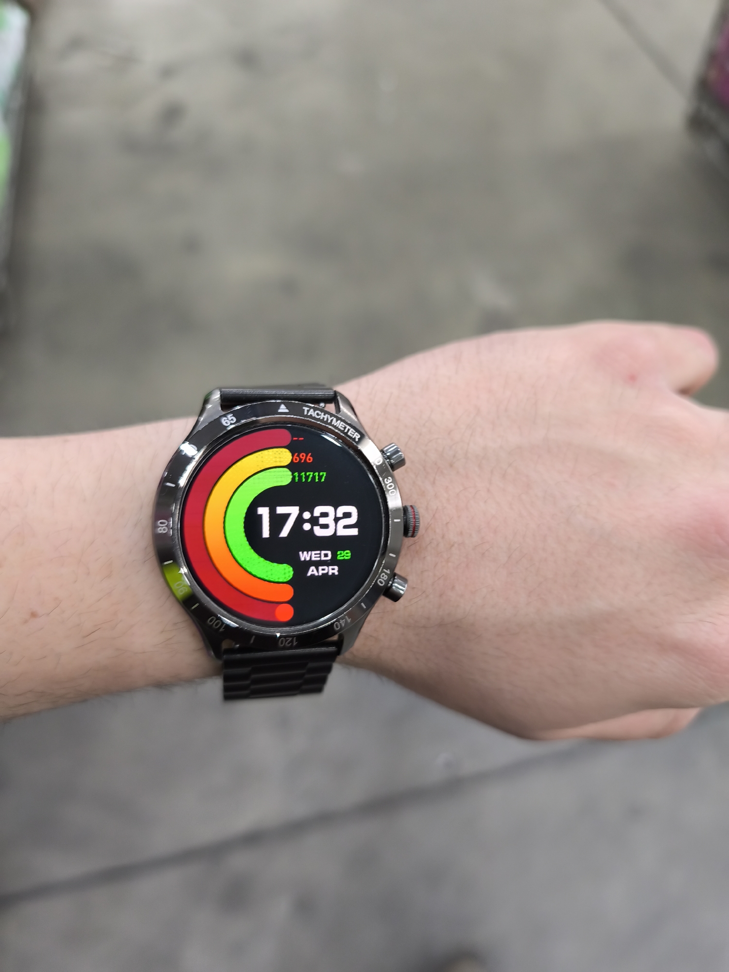 A close-up, slightly high-angle shot captures a person's left arm and hand, prominently featuring a smartwatch on the wrist. The background is intentionally blurred, revealing a grey, possibly concrete or tiled floor, indicative of an indoor environment like a store or public building. Hints of other blurry objects, including a green and white item on the far left, are visible in the background.
The smartwatch has a sleek black metallic case and strap. Its round digital display is brightly lit, showcasing various functionalities. The watch bezel is marked with "TACHYMETER" and numerous values (65, 80, 100, 120, 140, 180, 300). The watch face displays the time as "17:32" in bold white digits. Below the time, the date is shown as "WED 29 APR." To the upper left of the time, two smaller numerical readouts appear: "696" in red and "11717" in green, likely representing activity or health metrics. These numbers are partially encircled by a vibrant, multi-colored arc, transitioning from red through orange and yellow to green, which appears to be a progress indicator. The arm is fair-skinned with light hair. The scene is static, focusing on the smartwatch and its display. While the image does not contain specific visual cues to confirm the location, it is understood to be taken in Burgas, Bulgaria.