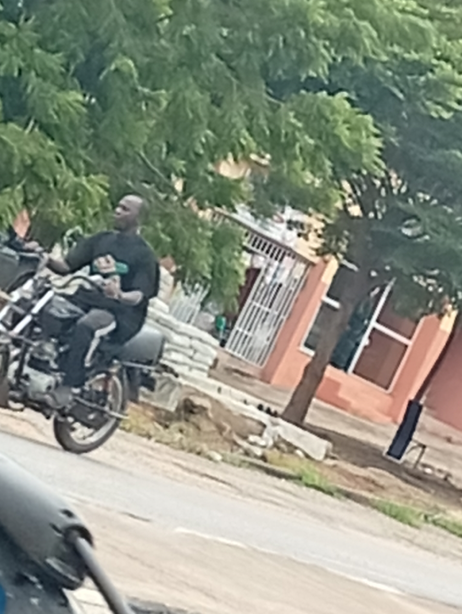 A man is riding a motorcycle on a paved road. The man is wearing a black t-shirt and black pants with a white stripe down the side. He is bald and appears to be looking to his left. The motorcycle is black and appears to be an older model. In the background, there is a building with orange walls and a white railing. There are also trees with green leaves. The road is gray and appears to be a two-lane road. There are some white sacks piled up on the side of the road.