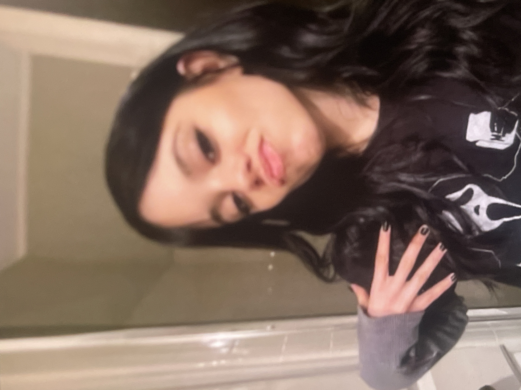 A rotated, blurry, close-up photograph captures a young person, likely female, with long, dark, wavy hair, from the chest up in an indoor setting in Poltava, Ukraine. The individual is positioned diagonally across the frame, with their head tilted. They are making a playful expression, sticking their tongue out slightly and pouting their lips. Their eyes appear dark and are looking towards the camera.
The person is wearing a dark, long-sleeved top or sweatshirt, which features a light-colored, abstract design on the right side of their chest/shoulder, resembling a skull or a stylized pattern. The cuff of the visible sleeve is grey and ribbed. A hand, adorned with dark nail polish, is raised with fingers spread, resting near their hair or shoulder.
The background is out of focus and indistinct, suggesting an interior space such as a bathroom or a room with light-colored walls or fixtures like a countertop. The lighting is soft and appears somewhat dim. No discernible text is visible in the image. The overall mood conveyed is casual and lighthearted.