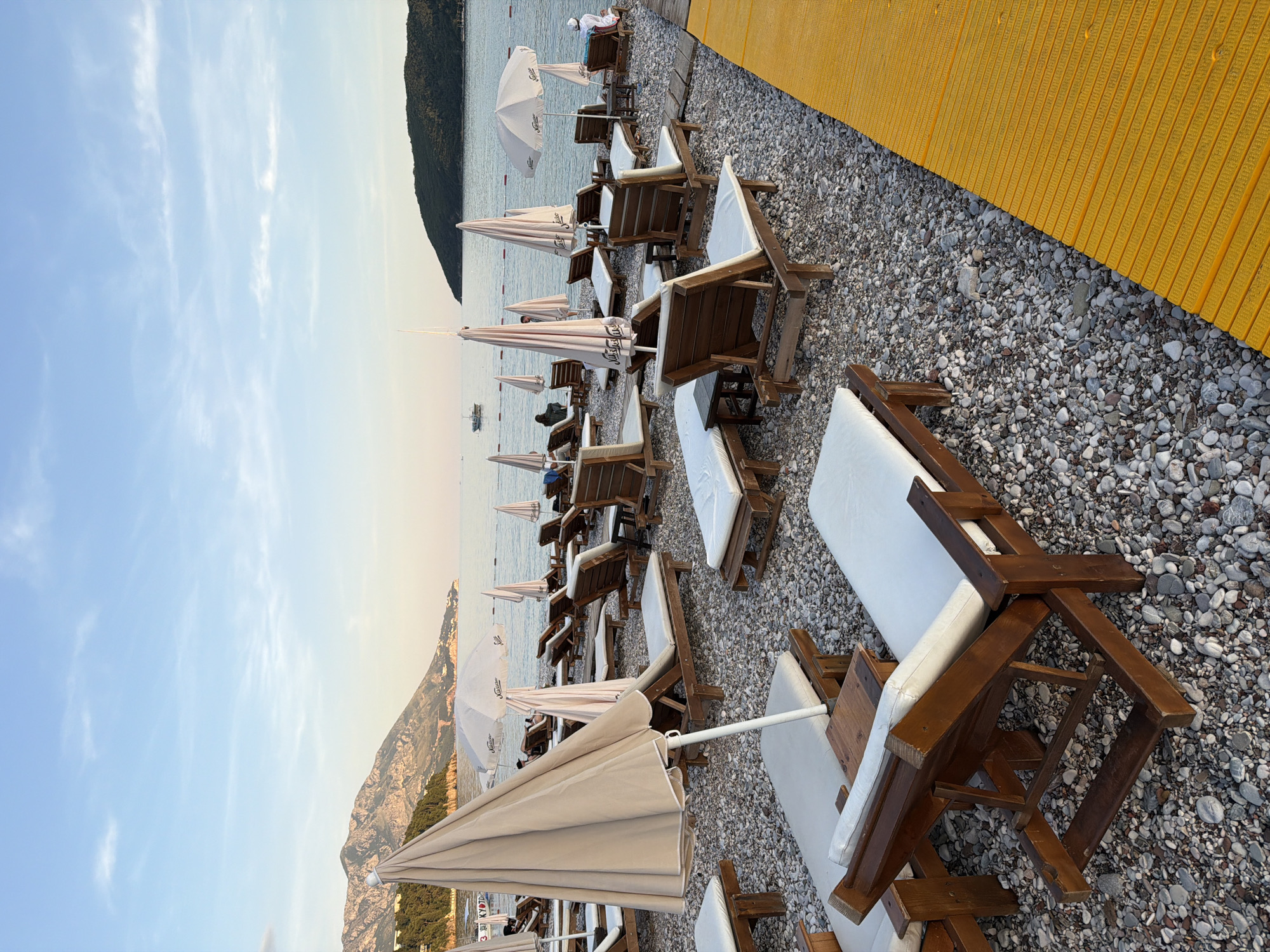The image captures a beach scene in Budva, Montenegro, with rows of wooden beach chairs topped with white cushions, arranged neatly under beige parasols. The beach is covered in small grey pebbles, and a yellow wooden boardwalk runs along the edge. The Adriatic Sea stretches out to the horizon, with a mountainous island visible in the distance. The sky is a pale blue, suggesting either early morning or late afternoon. No specific individuals or actions are emphasized; the scene is a general view of a prepared beach setting.