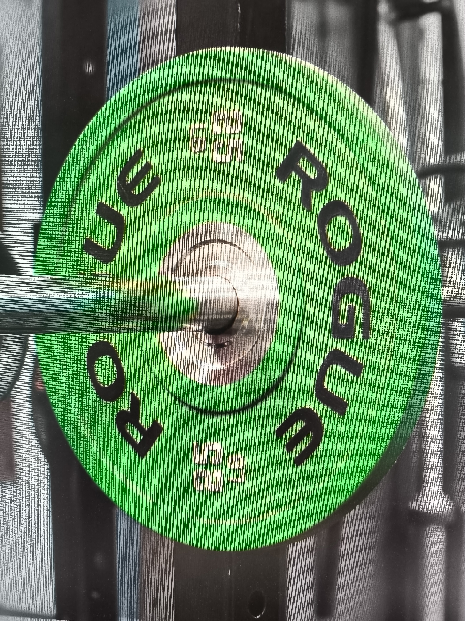 A green weight plate with black lettering is on a silver barbell. The plate has "ROGUE" written in a circular pattern on its surface. To the left of "ROGUE" there are some markings in what appear to be Russian and numbers. The weight plate has a textured surface. The background is out of focus and appears to be a gym setting with other weight plates and equipment. There are no people present in the image. The location in the city of Sysert, Russia is unknown.