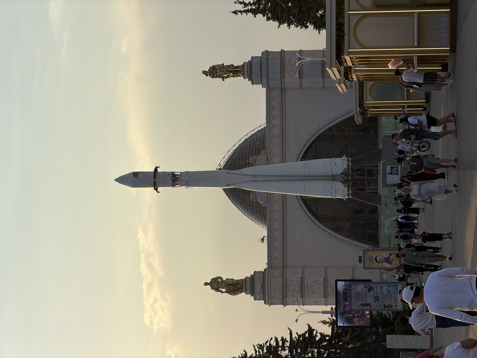 The image depicts a public space in Moscow, Russia, featuring a large white rocket standing vertically. The rocket is situated in front of a grand, ornate building with a glass dome and sculptures on its roof. A crowd of people is walking along a pathway in front of the building. Some people are standing near a concession stand selling pink cotton candy. Two large digital advertising panels are visible near the bottom of the frame. The sky is a pale gradient color, suggesting the time of day is either early morning or late afternoon.