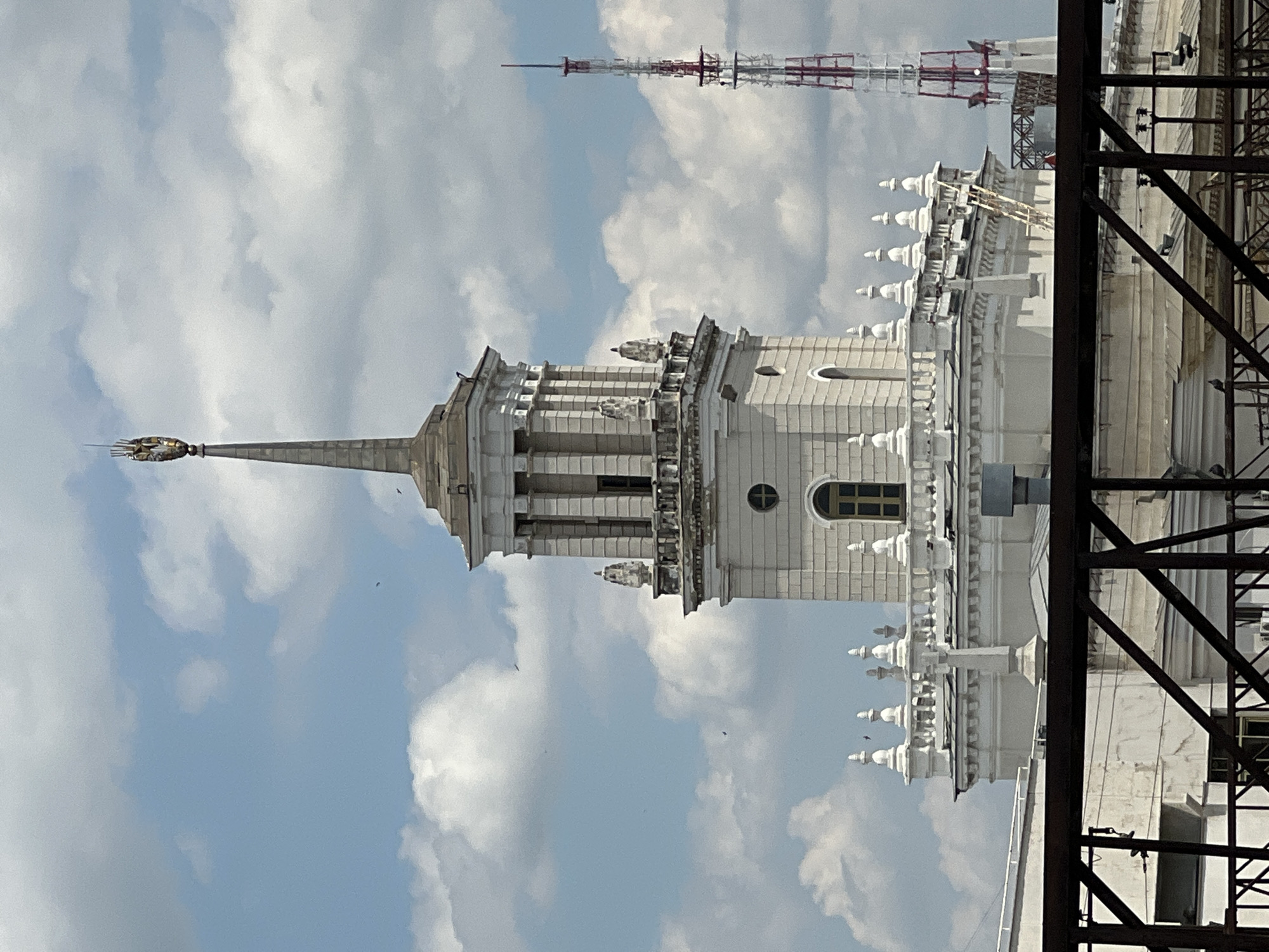 The image depicts a view of the upper sections of a white, ornate building, possibly a Stalinist Empire style skyscraper, against a bright blue sky filled with fluffy white clouds. The building features a central spire topped with a golden finial that appears to be a stylized winged object. The spire is surrounded by intricate architectural elements, including columns, decorative balustrades, and smaller domed structures. The building's white facade contrasts with the blue sky.
In the background, a tall, red and white lattice transmission tower is visible. On the right side of the image, a dark metal structure, possibly scaffolding or part of a larger construction, partially obscures the view of the building.
The lighting suggests it is daytime, likely midday or early afternoon, given the brightness of the sky and the clarity of the clouds. The weather appears to be pleasant, with clear skies and scattered clouds. There are no people visible in the image. The scene evokes a sense of grandeur and architectural detail. No text is visible in the image. The location is identified as Krasnodar, Russia, which is consistent with the architectural style and the presence of such transmission towers.