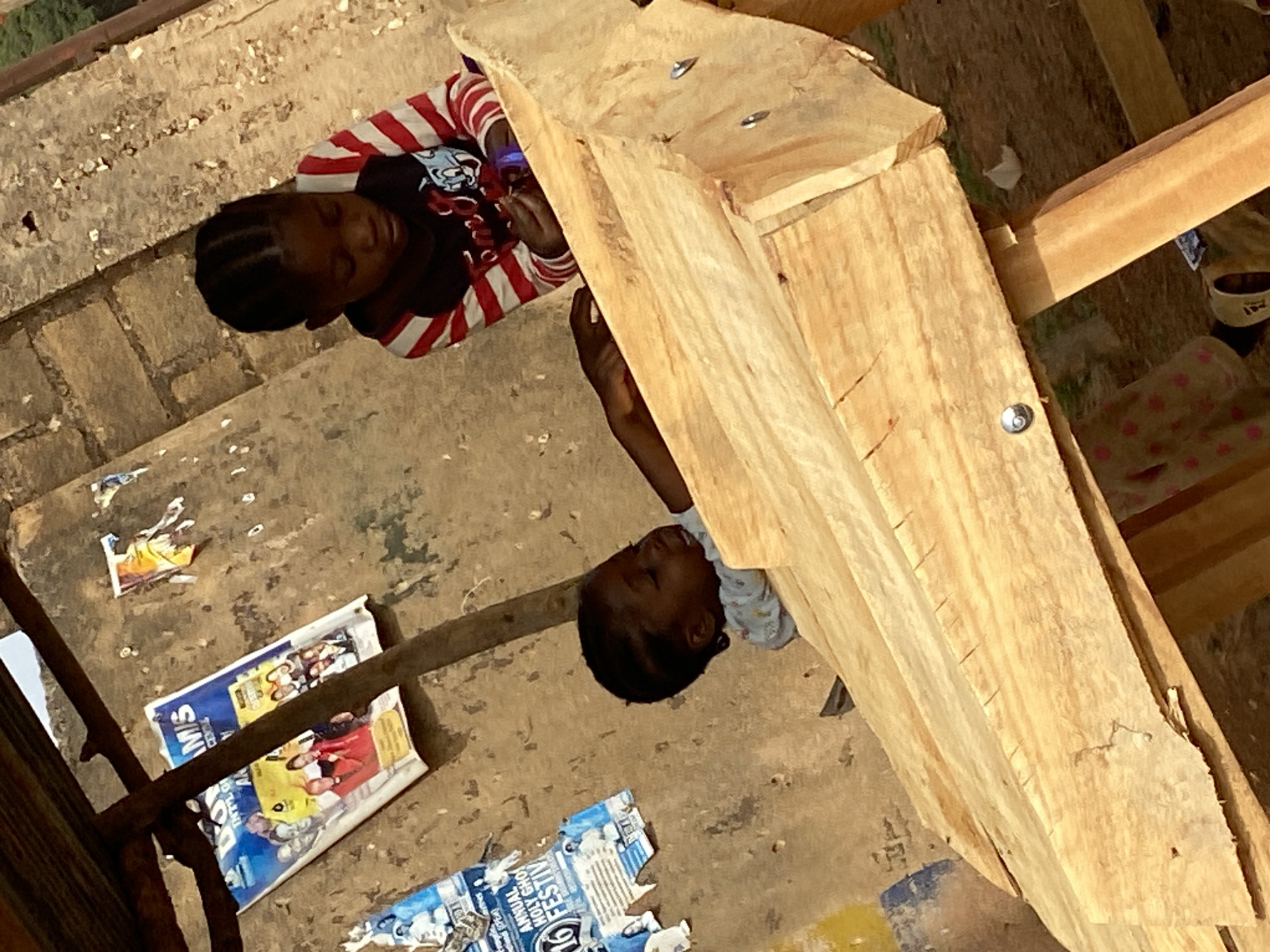 This image, captured in Bukuru, Nigeria, depicts two young children resting in a rustic outdoor setting dominated by wooden structures. The scene is bathed in warm, direct daylight, suggesting a sunny day.
In the foreground, one child with dark skin and braided hair is visible, wearing a red and white striped long-sleeved shirt over a dark t-shirt featuring a graphic. His eyes are closed or heavily lidded, indicating he is sleeping or very drowsy. His head is positioned beneath a substantial wooden beam. To the right and partially obscured by the wooden framework, a second child, also with dark skin and braided hair, is visible. This child appears to be wearing a light-colored, possibly patterned, long-sleeved top and is similarly at rest, with their eyes closed or nearly closed.
The children are positioned on an uneven dirt or concrete ground, which is strewn with various pieces of litter, including torn paper and flyers. Visible text fragments from these papers include "ANNUAL HOLY GHO FESTIV" and "INT'L GI", likely from local event advertisements. The wooden structures, appearing to be rough-hewn timber planks joined by screws or bolts, suggest either a construction site or a makeshift shelter. The overall impression is one of peaceful slumber in a simple, utilitarian environment.