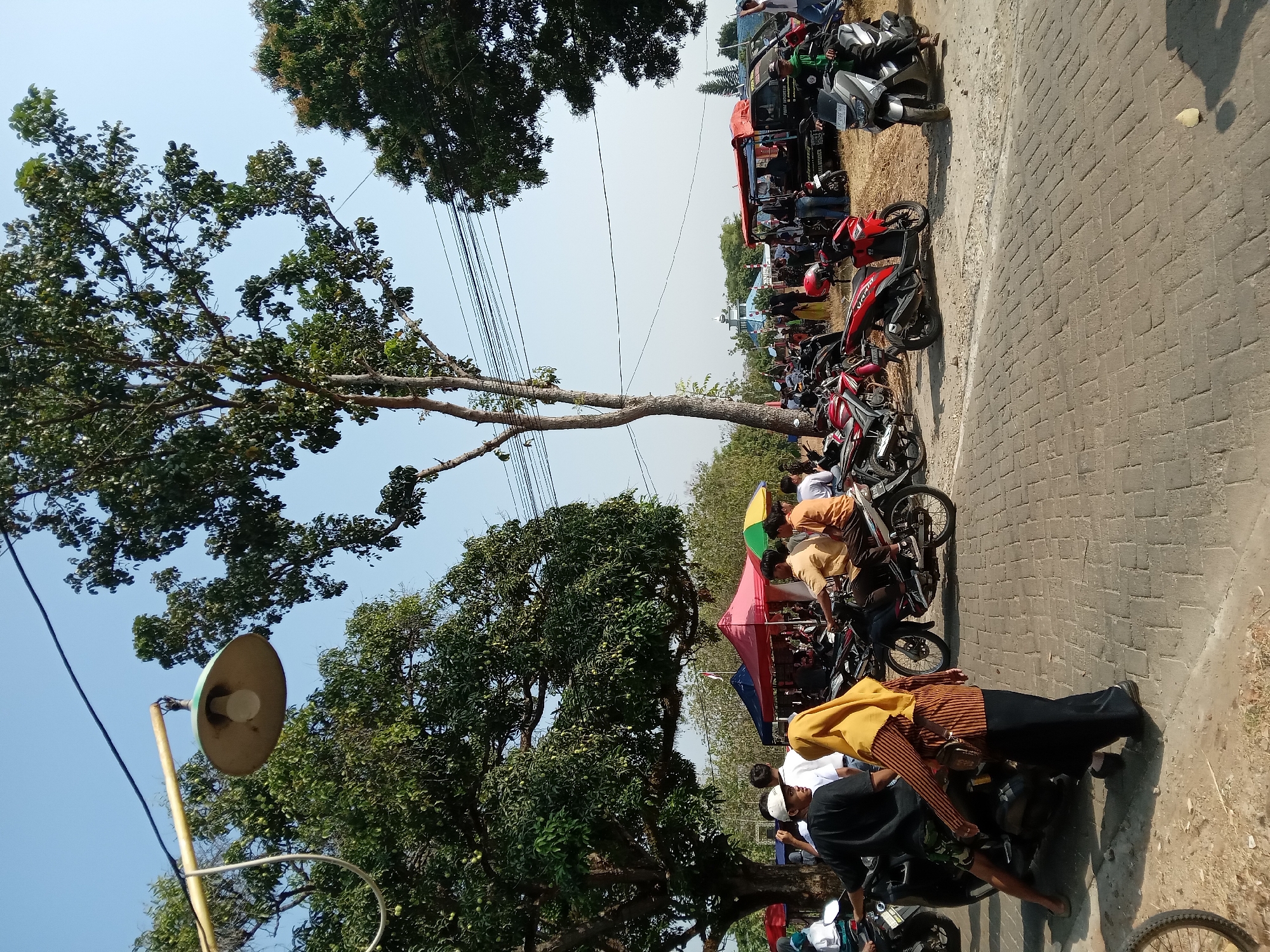 The image depicts an outdoor scene in Ngrambe, Indonesia, on a clear, sunny day. The foreground features a paved area with several motorcycles parked, and a dirt path to the left. In the middle ground, a group of people are gathered around tents, which appear to be stalls or shelters. Some individuals are on motorcycles, while others are walking or standing. The background reveals trees, power lines, and a distant building with a light blue roof and steeple, possibly a mosque. The overall atmosphere suggests a community gathering or market event.