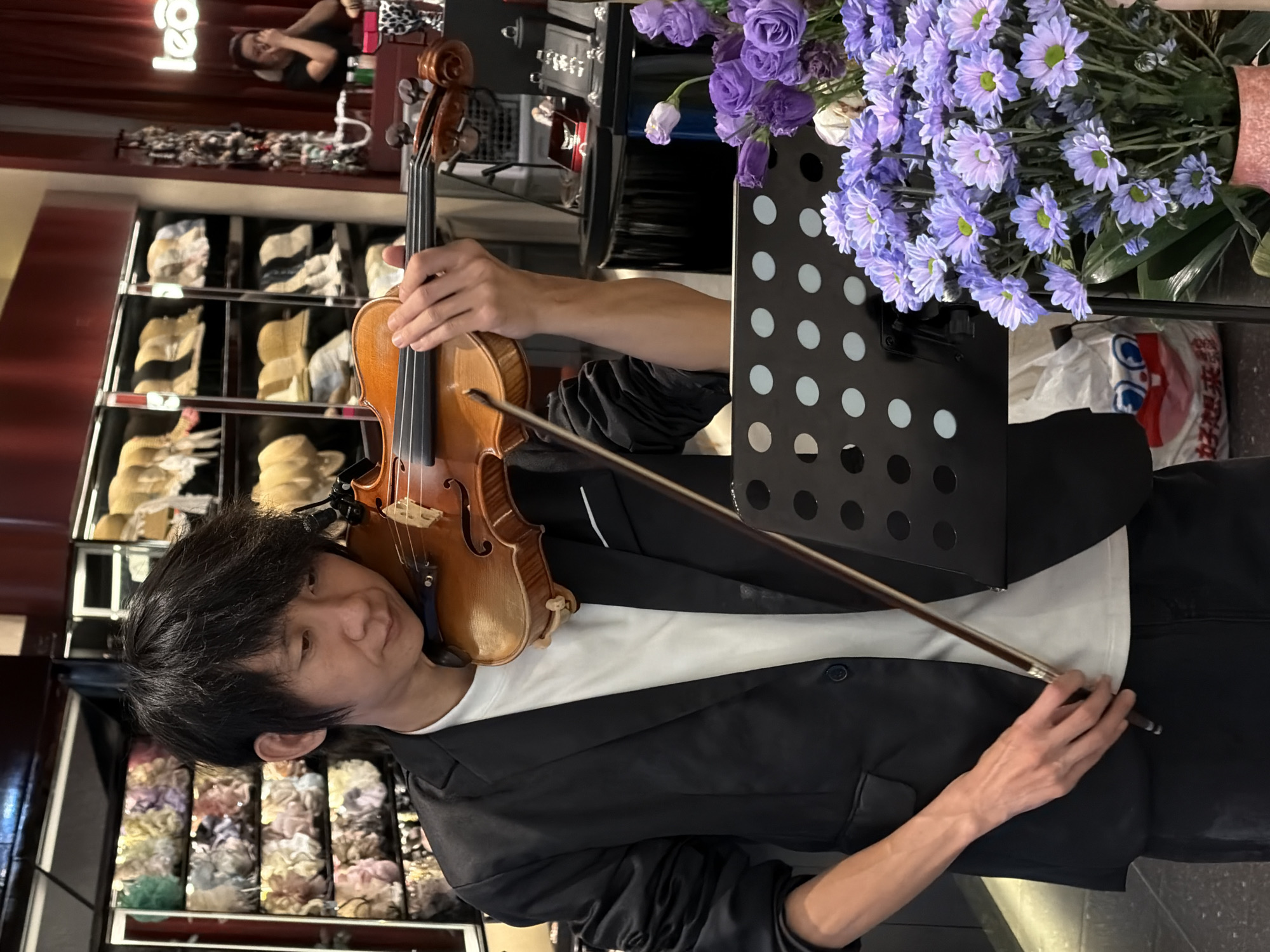 An East Asian male musician is depicted playing a violin in a retail store located in Yuzhong District, China. The man, positioned slightly to the left of the frame's center, wears a black blazer over a white t-shirt and dark pants. His dark hair is styled to frame his face, and his gaze is directed downwards towards his instrument and a black perforated music stand positioned in front of him. He holds a light brown wooden violin against his left shoulder, with his left hand's fingers placed on the strings near the scroll. His right hand grips a dark bow with a silver-colored ferrule, actively drawing it across the violin's strings.
The background reveals a well-lit retail environment. To the man's left and right are tall display shelves. The shelves on the left contain neatly arranged rows of soft, fluffy items, possibly hair accessories or textile products, in various pastel shades including green, yellow, pink, and blue. To the right, shelves showcase numerous hats, predominantly straw hats and bucket hats, in natural and light tones like beige and white. Further in the background, on the far right, a red wall features a white illuminated sign that reads "le&co". Below this sign, a woman with dark hair in a bun and a dark top is visible behind a counter, hands raised near her face. Jewelry displays, including rings and watches, are visible on dark stands on the counter to the right.
In the bottom right foreground, a vibrant bouquet of fresh flowers is displayed. The bouquet consists primarily of light purple chrysanthemums with prominent green centers, along with some darker purple roses or similar flowers. These are arranged in a pink or terracotta-colored vase. Partially obscured behind the flowers and the music stand is a white plastic shopping bag featuring a red and blue cartoon face and Chinese characters that translate to "really want a pack." The floor appears to be a dark, polished surface.