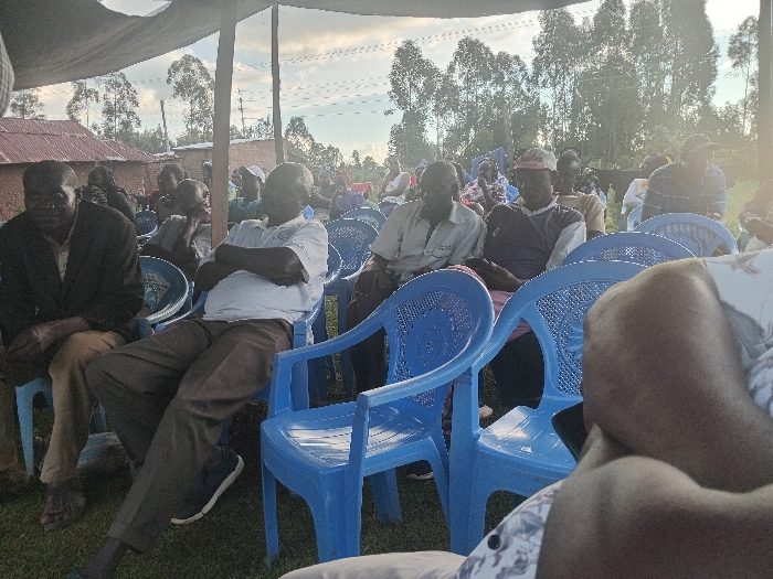 The image depicts an outdoor gathering in Chevaywa ward, Kenya. Numerous individuals, predominantly adult males, are seated on blue plastic chairs under a dark canopy or tent. In the foreground, the legs and lap of a person in patterned shorts are visible, holding a dark-colored mobile phone. To the left, a man in a dark jacket and light shirt sits, followed by another man in a white polo shirt and dark trousers with his arms crossed. Further back, additional attendees are visible, some wearing hats, all seated and facing generally forward, suggesting an organized meeting or event. The background features rural structures with brick/mud walls and corrugated iron roofs, backed by a line of tall, slender trees and overhead utility lines. The ground is covered with grass, and the sky is partly cloudy with a bright, possibly late-day, atmospheric lighting.