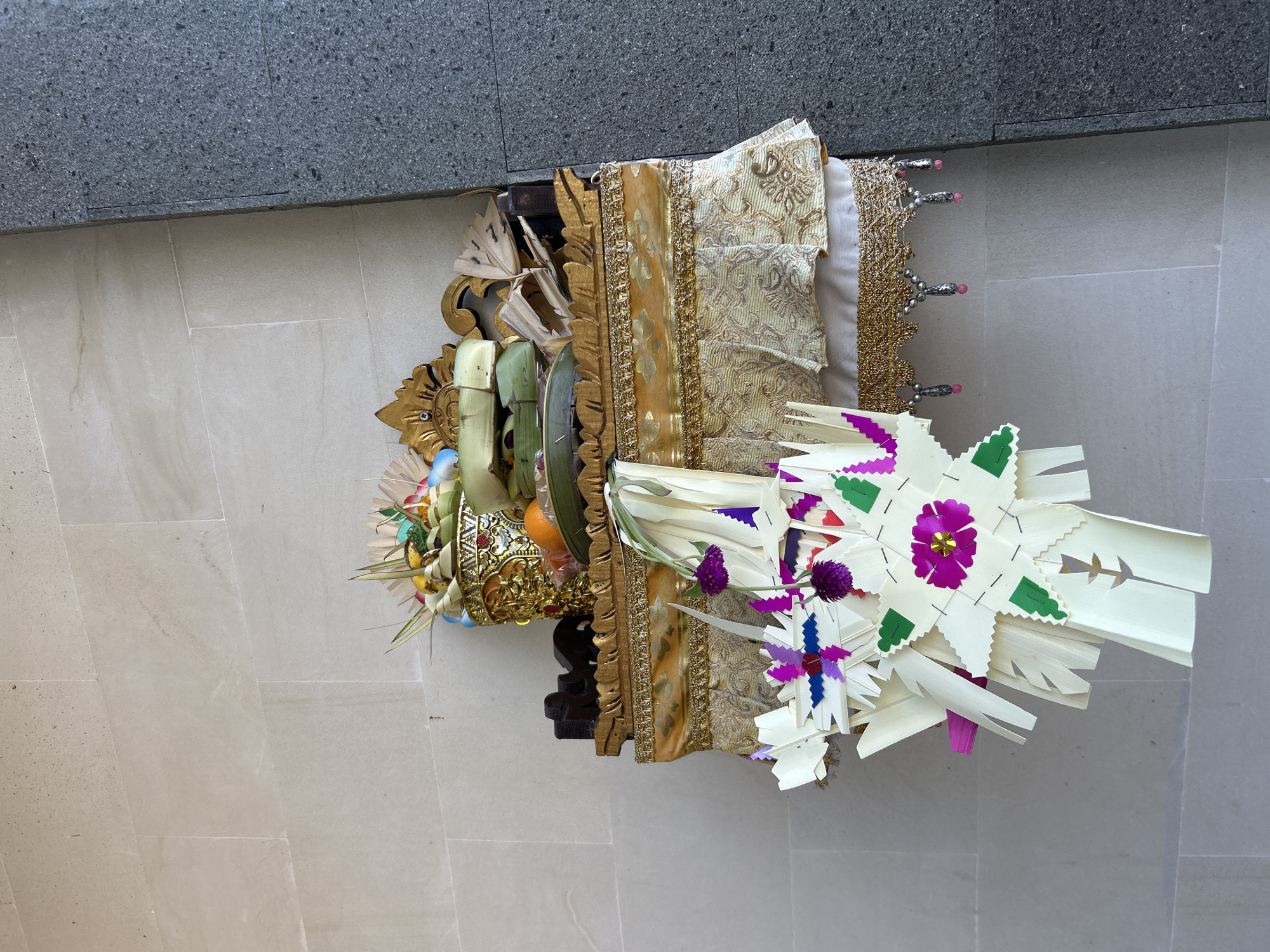 The image displays a Balinese offering, likely a Canang Sari, presented on a decorative stand. The offering is composed of various natural elements, including fruits, flowers, and intricately folded palm leaves. The palm leaves are fashioned into star-like shapes and adorned with colorful paper or flower petals in shades of purple and green. The stand itself is ornate, featuring gold-colored detailing and fabric adornments.
The scene appears to be outdoors, suggested by the lighting and the tiled ground, which resembles an outdoor patio or walkway. A gray, textured wall forms the backdrop at the top of the image, possibly indicating an urban or developed area. The lighting suggests it is daytime, with no specific indications of weather conditions. The overall impression is one of religious or cultural significance, given the nature of the offering. There is no visible text in the image.