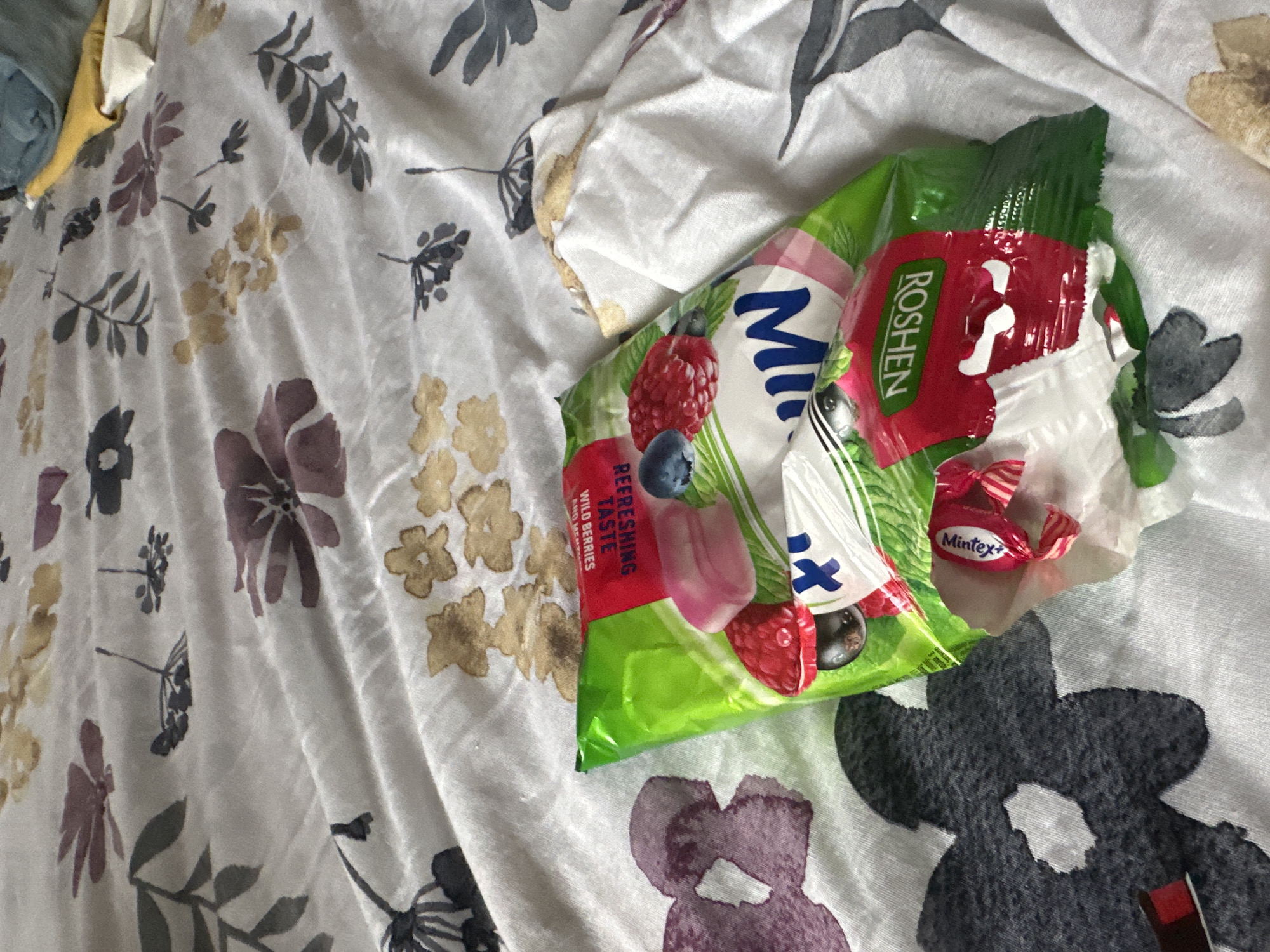 A close-up, slightly overhead shot shows a packet of Roshen Mintx candies resting on a white duvet cover with a floral pattern. The duvet cover features scattered illustrations of various flowers and leaves in muted colors, including purples, yellows, and grays, with some darker, abstract floral shapes towards the bottom right. The packet of candies is green and white with colorful fruit illustrations, including raspberries and blueberries. Text on the packet reads "Roshen" in a red rectangular box and "Mintx+" on a small, pink wrapped candy beside it. Another line of text on the packet says "REFRESHING TASTE WILD BERRIES." The lighting suggests it is daytime, with soft, diffused light. The overall scene is casual and domestic, possibly a bedroom. The image context places the scene in Budva, Montenegro.
