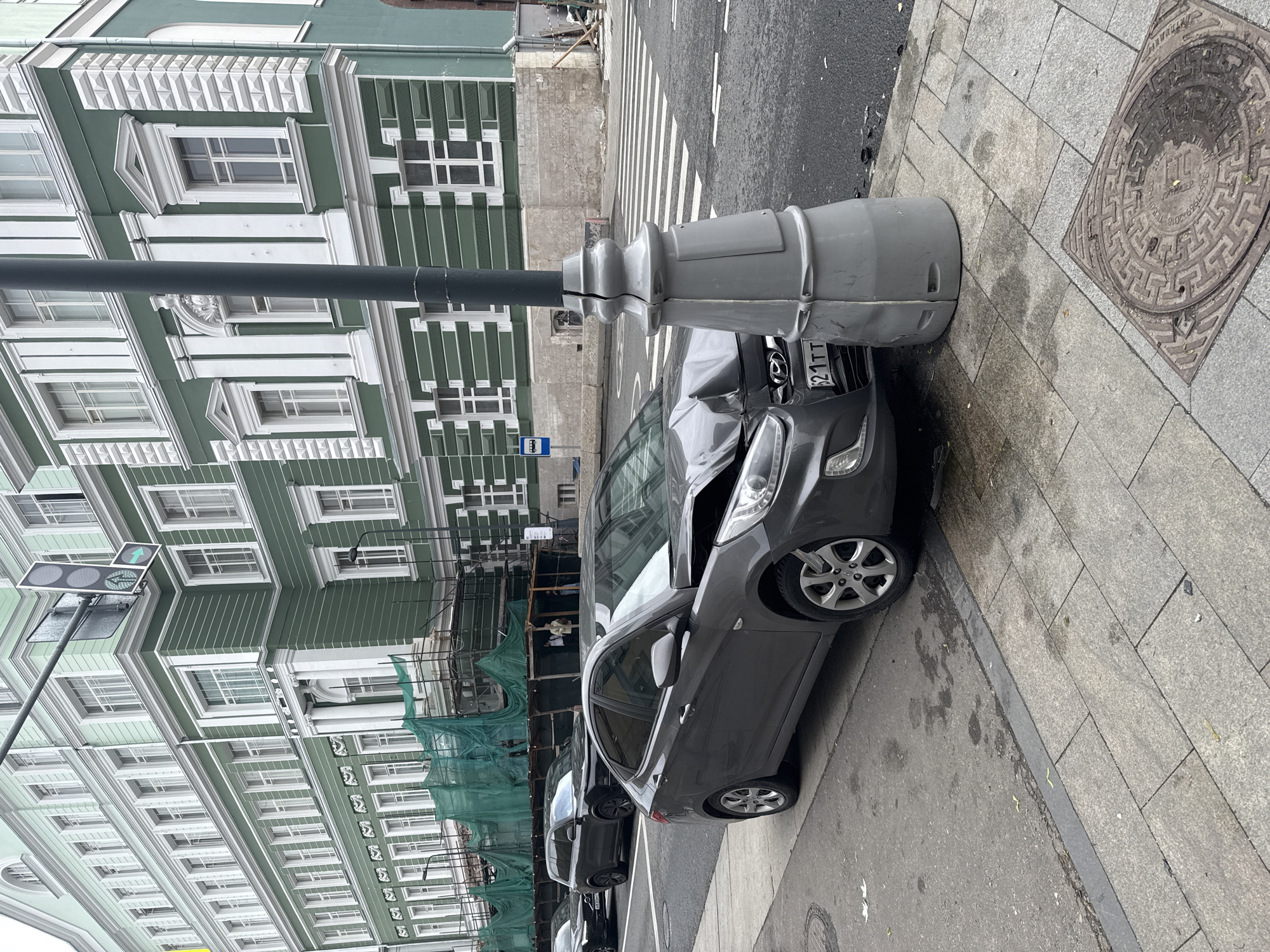 The image depicts a car accident scene in Moscow, Russia. A gray Hyundai Accent is severely damaged, with its front end crushed against a gray lamppost on a sidewalk. The car's hood is buckled, and the windshield is cracked. The lamppost is situated near a manhole cover on the sidewalk. In the background, there is a building with a green facade and multiple windows. A traffic light is visible above the street. Scaffolding is set up on the building, indicating construction or renovation work. Other cars are parked along the street behind the damaged vehicle. A bus stop sign is also visible.
