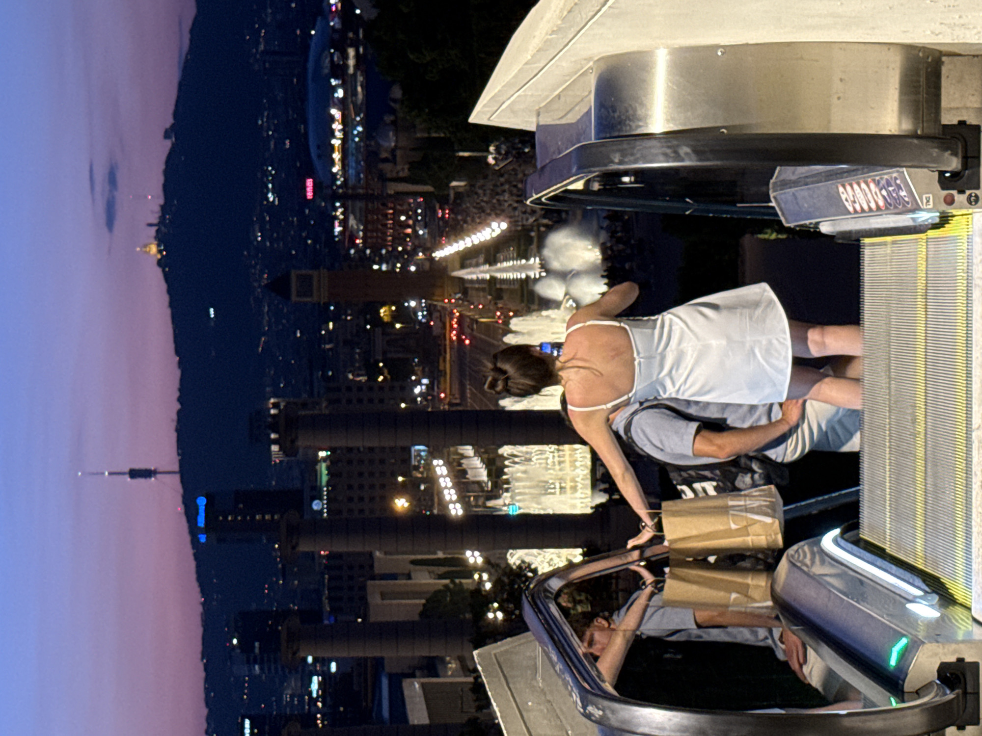 The image depicts an outdoor escalator at dusk, overlooking the city of Barcelona, Spain. The cityscape in the background is illuminated by city lights and features a prominent hill topped with structures and a communication tower. In the mid-ground, a series of fountains are active, and a wide avenue is lined with lights and traffic. On the escalator, two individuals are visible from behind. One, wearing a white dress, is standing and appears to be taking a photo with a smartphone. To their left, another person, possibly a male, is holding two brown paper shopping bags. The escalator itself is metallic with yellow safety lines on the steps. The sky above transitions from a deep blue at the top to a soft purple and pink near the horizon.