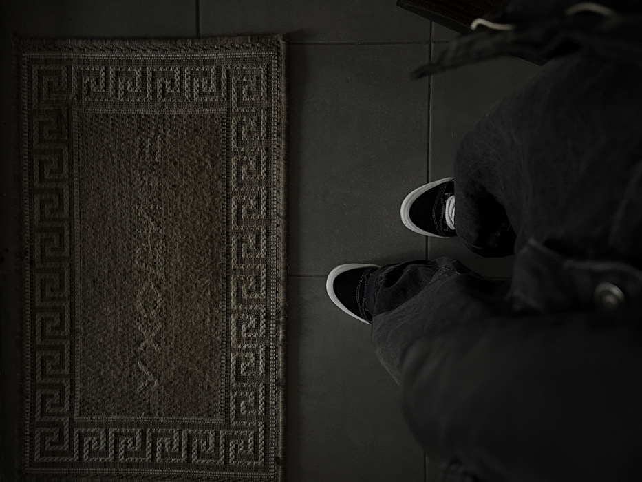 The image presents an overhead, first-person view depicting a person's lower legs and feet positioned on a dark tiled floor. The individual is wearing dark, loose-fitting trousers and black sneakers with white soles and toe caps. Directly in front of the feet lies a rectangular, dark brown doormat. This mat features a distinct woven Greek key (meander) pattern border. The Cyrillic word "УЮТНО" (meaning "cozy" or "comfortably") is visibly embossed or woven into the center of the mat. The floor consists of dark grey square tiles with clearly defined grout lines. The scene depicts an indoor entrance area, likely a hallway or vestibule, located within the city of Moscow, Russia, as evidenced by the Russian text on the doormat.